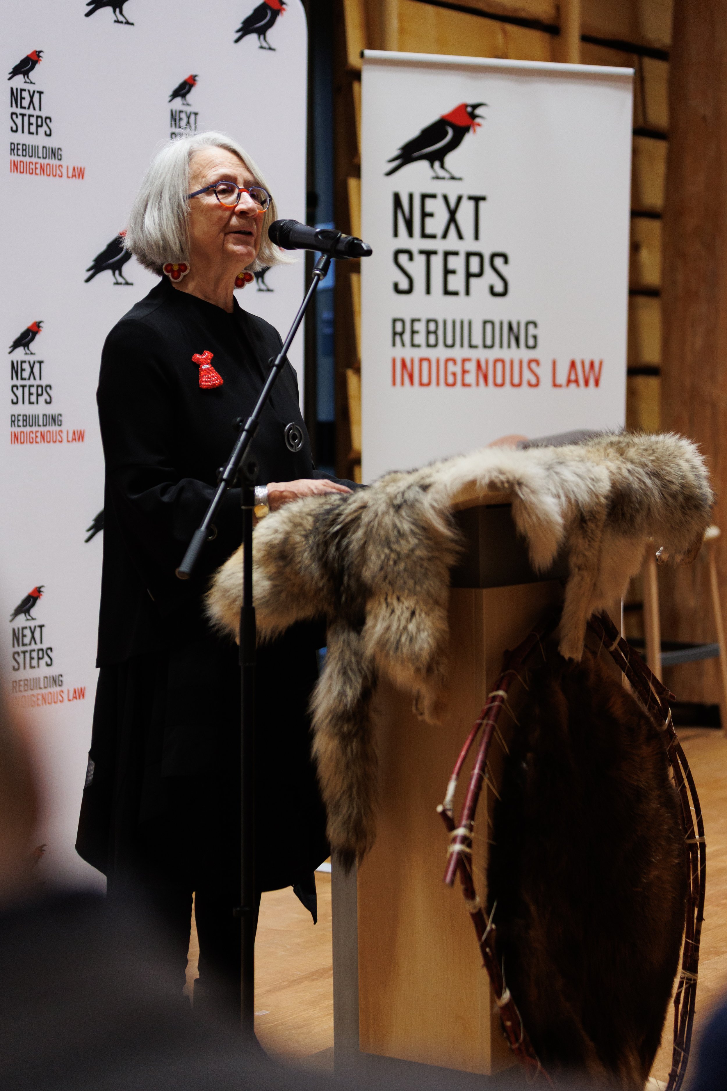 An older woman with gray hair and glasses speaking at a podium decorated with fur, in front of two banners that read "NEXT STEPS REBUILDING INDIGENOUS LAW" and have a black bird with red markings.