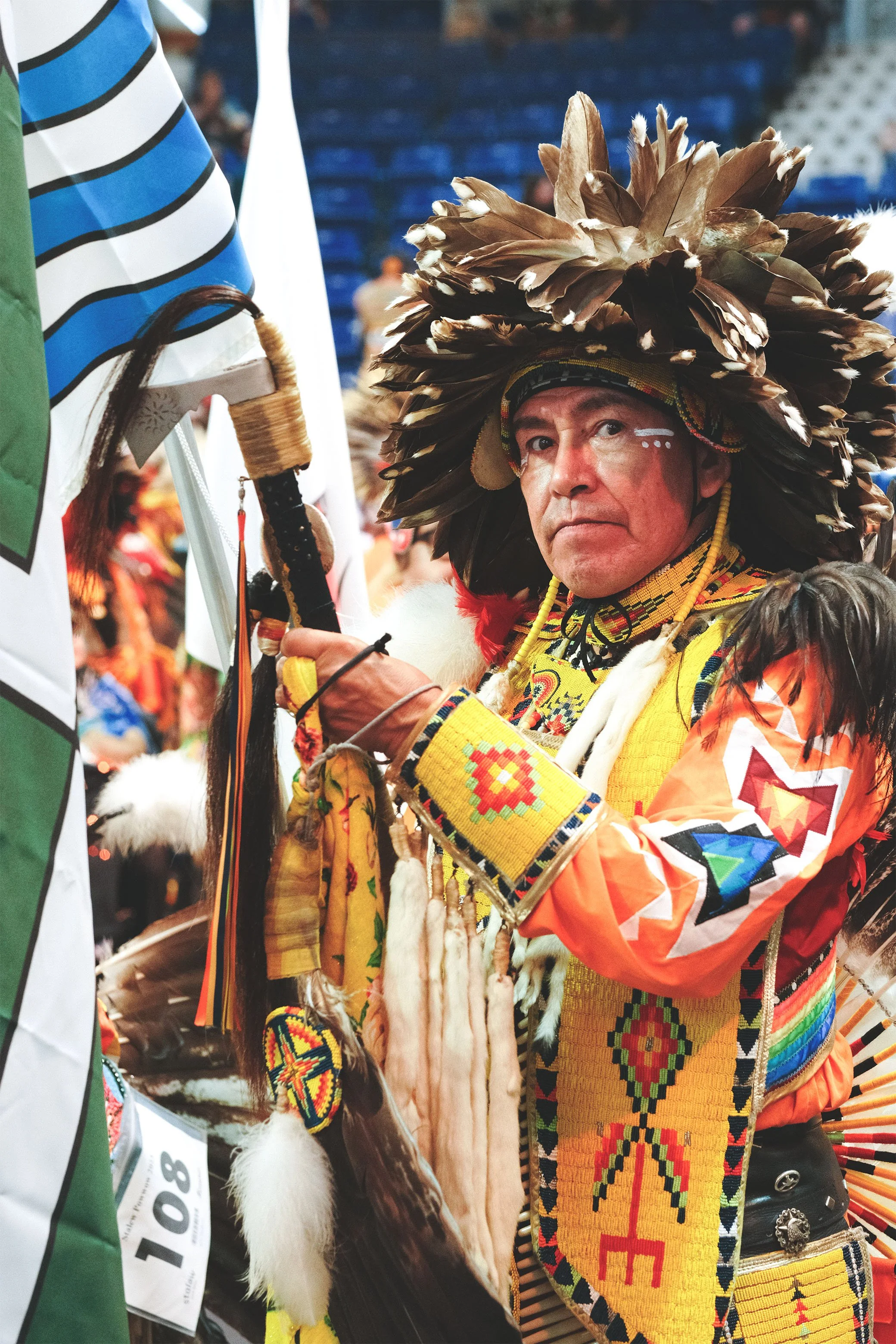 A Indigenous man dressed in traditional attire with a feathered headdress, holding a staff at a cultural event.