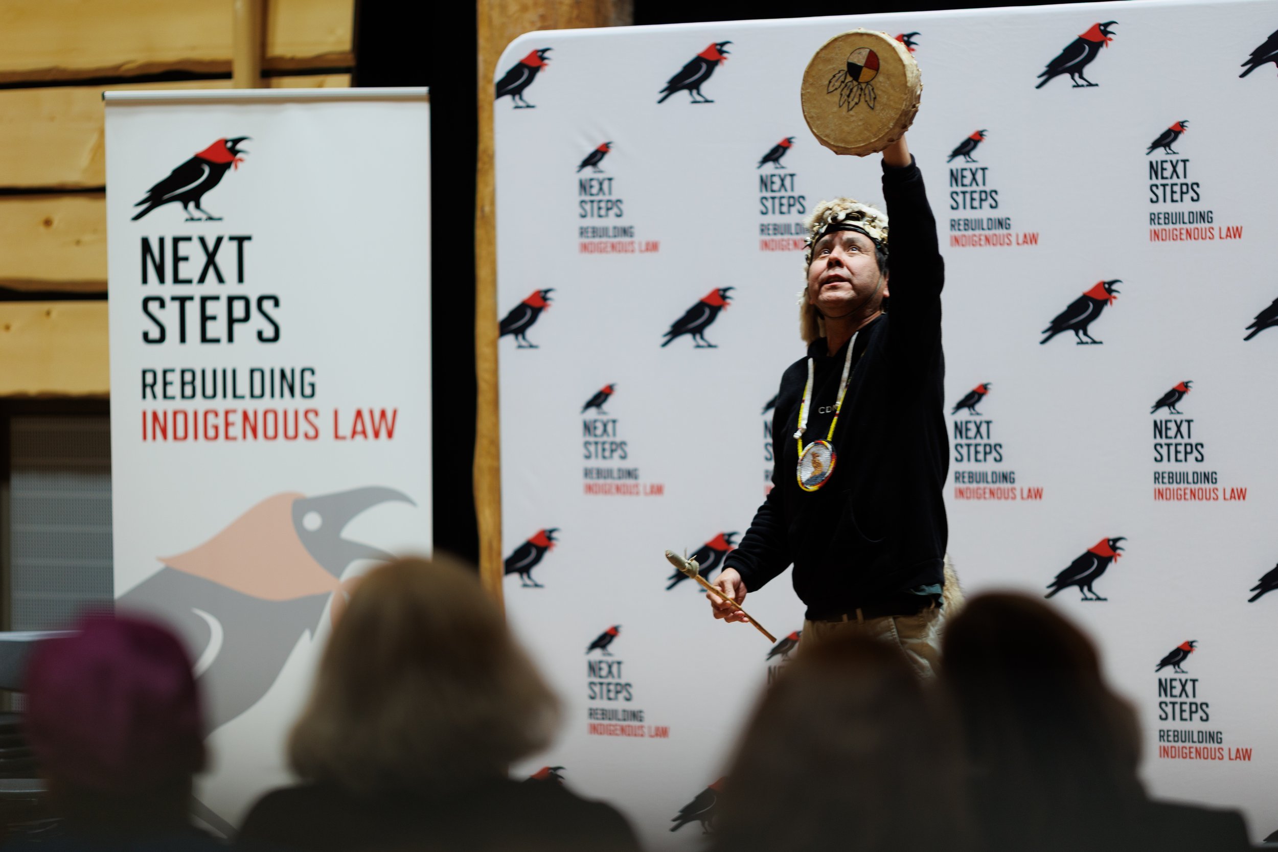 A person dressed in traditional Indigenous attire performs a dance with a circular drum at a conference titled 'Next Steps Rebuilding Indigenous Law', standing in front of a backdrop and banner featuring a black  with a red crest and the event's slogan.