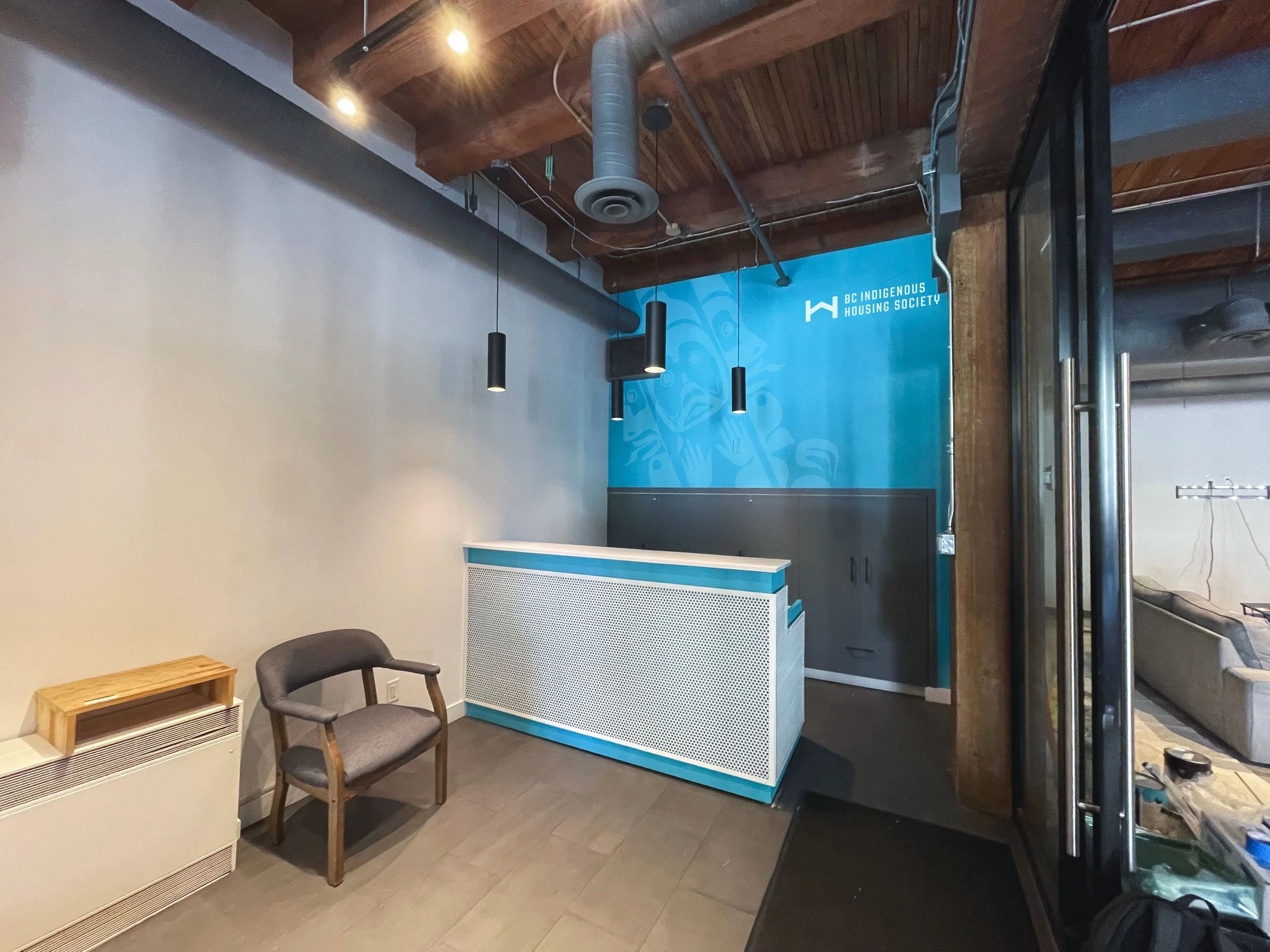 Reception area with a wooden chair, a small white heater, and a reception desk with a blue top. Behind the desk is a blue wall with the BC Indigenous Housing Society logo. The ceiling has exposed wooden beams and hanging black light fixtures.