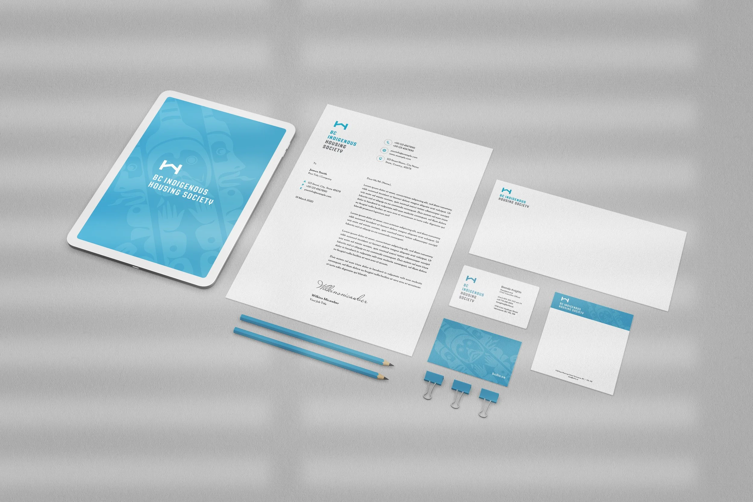 Collection of office stationery items for BC Indigenous Housing Society, including a tablet with logo, letterhead, business cards, envelope, folder, pencils, paper clips, and binders, all featuring the organization's blue and white branding.