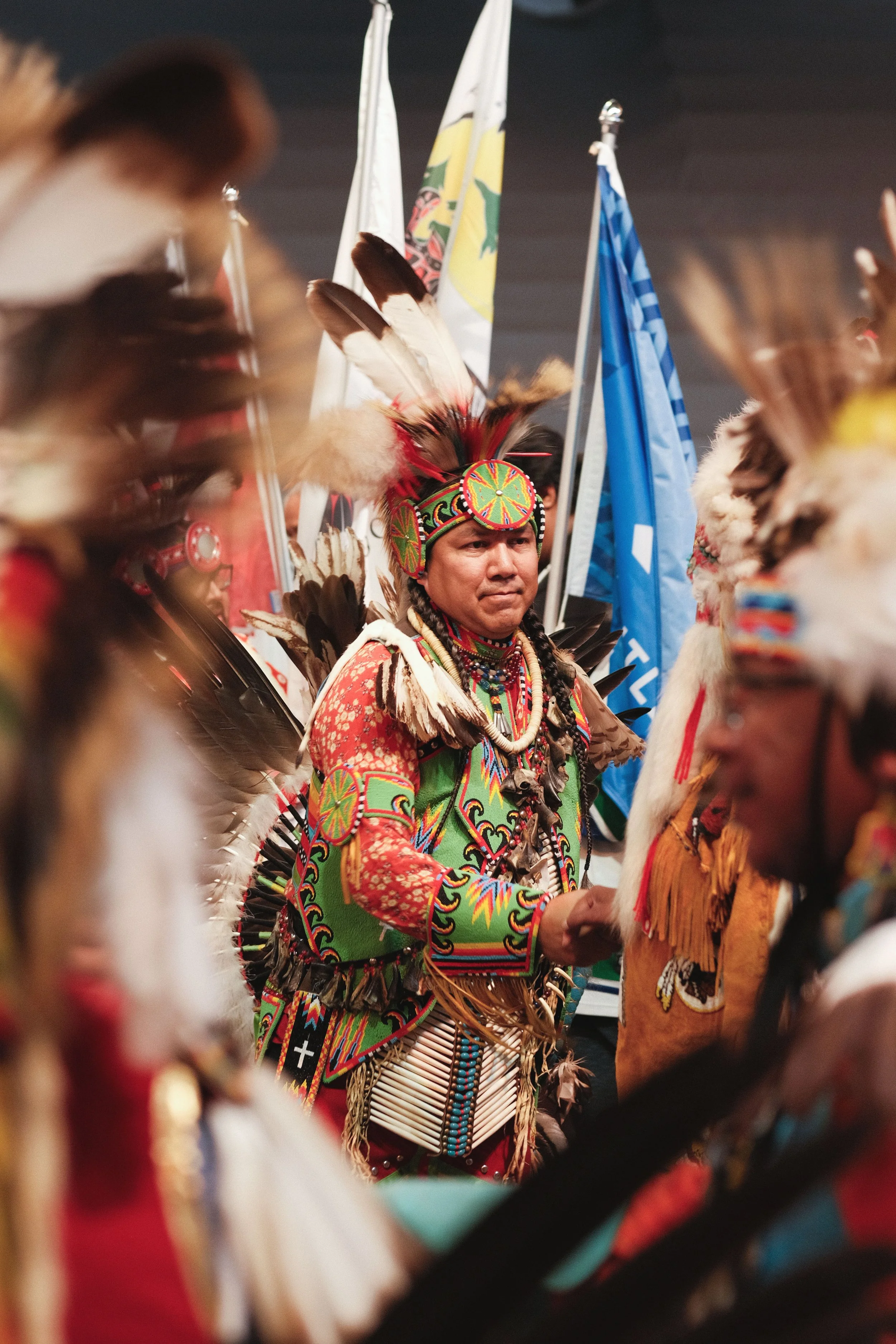 A Native American man wearing traditional regalia with feathers, beads, and colorful embroidered clothing, participating in a cultural ceremony or gathering, with flags in the background.