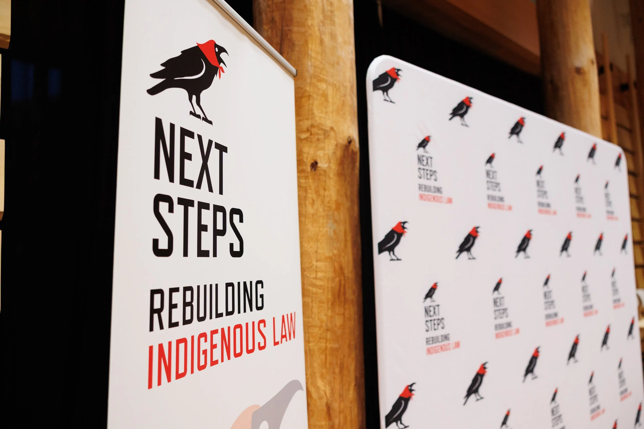 Signs with a black raven logo, the text 'Next Steps Rebuilding Indigenous Law,' and repeated crow logo pattern on a white background, wooden wall background.