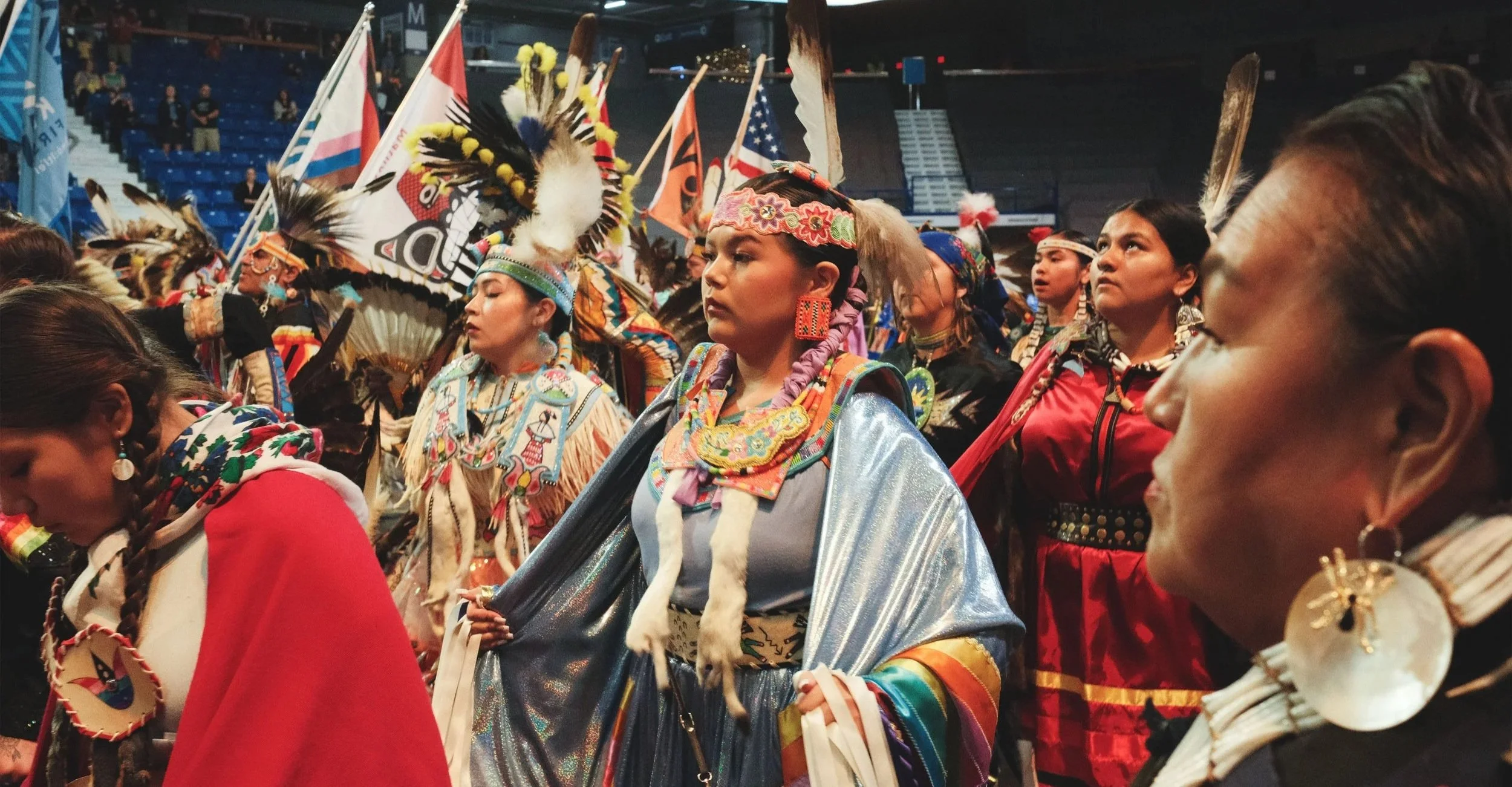 Native American women and young girls dressed in traditional regalia, including feathered headdresses, beaded jewelry, and colorful clothing, participating in a cultural ceremony or event inside an arena with flags and spectators in the background.