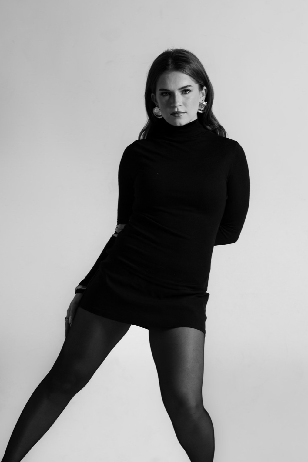 Black and white photo of a woman in a black dress and tights, posing with one leg forward, looking at the camera.