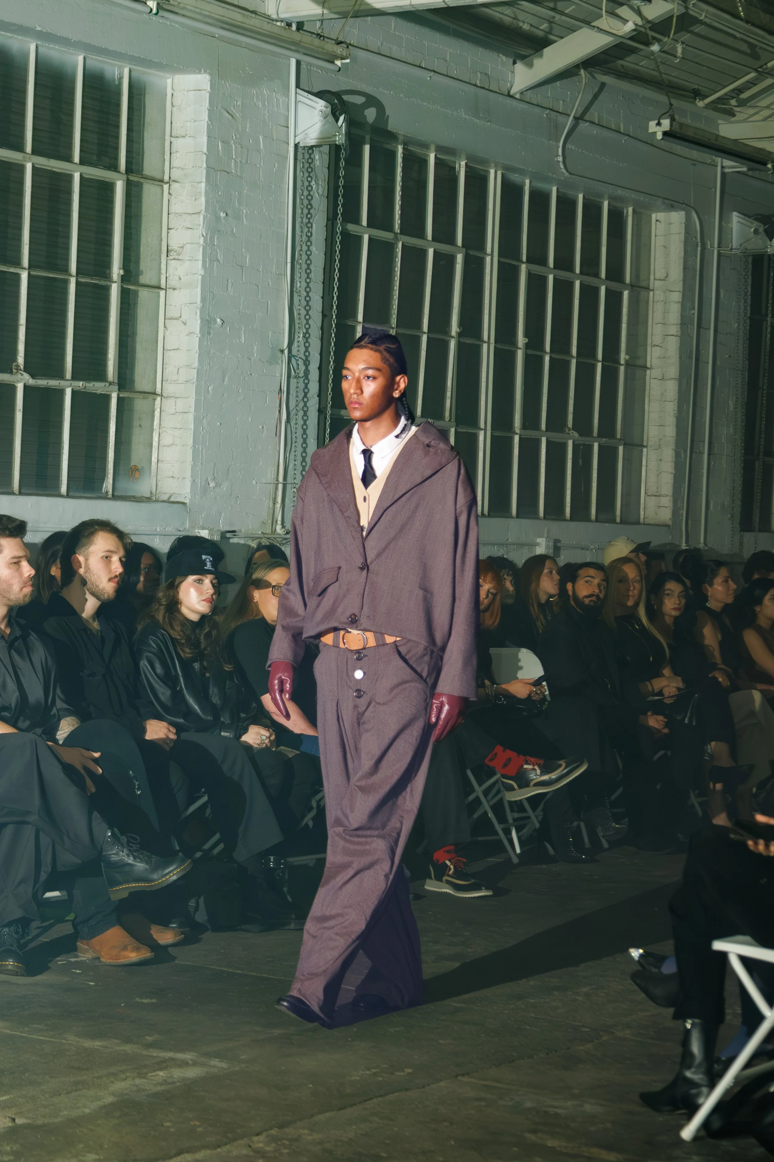Model walking down the runway during a fashion show in an industrial space with a gray brick wall and large windows.