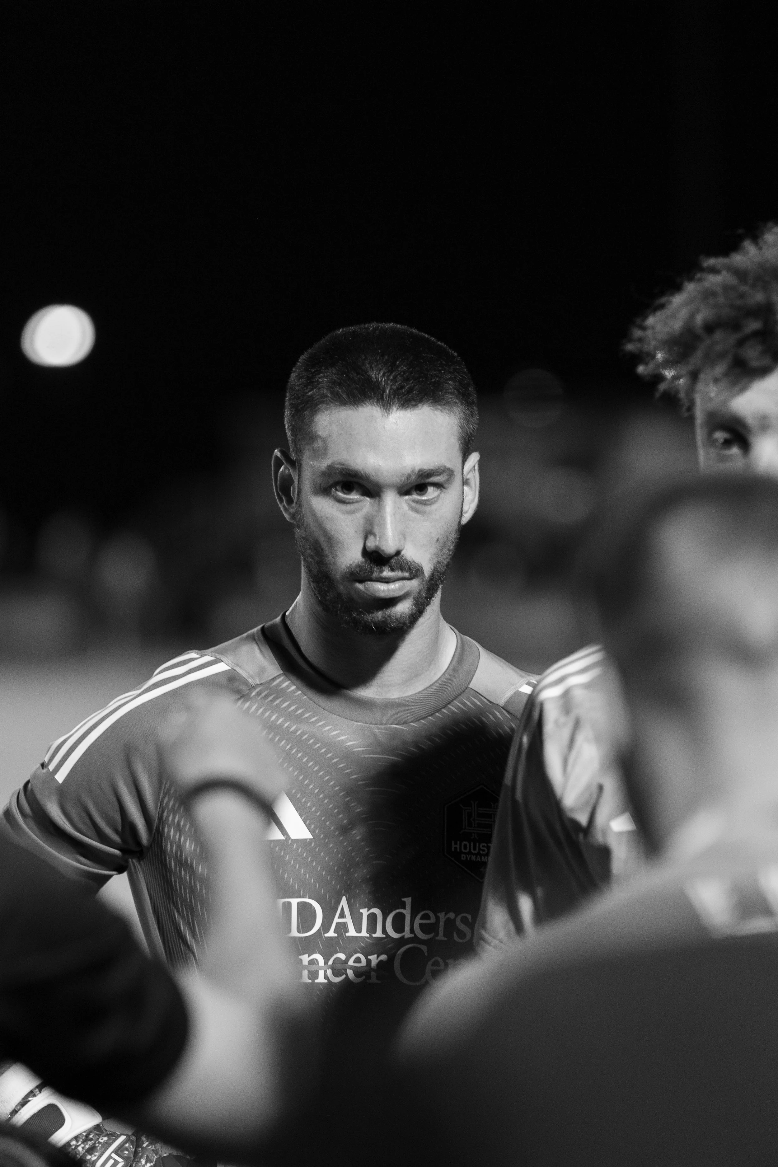 A black and white photograph of a man with short dark hair and a beard wearing a sports jersey, looking directly at the camera with a serious expression, surrounded by other players with blurred faces.