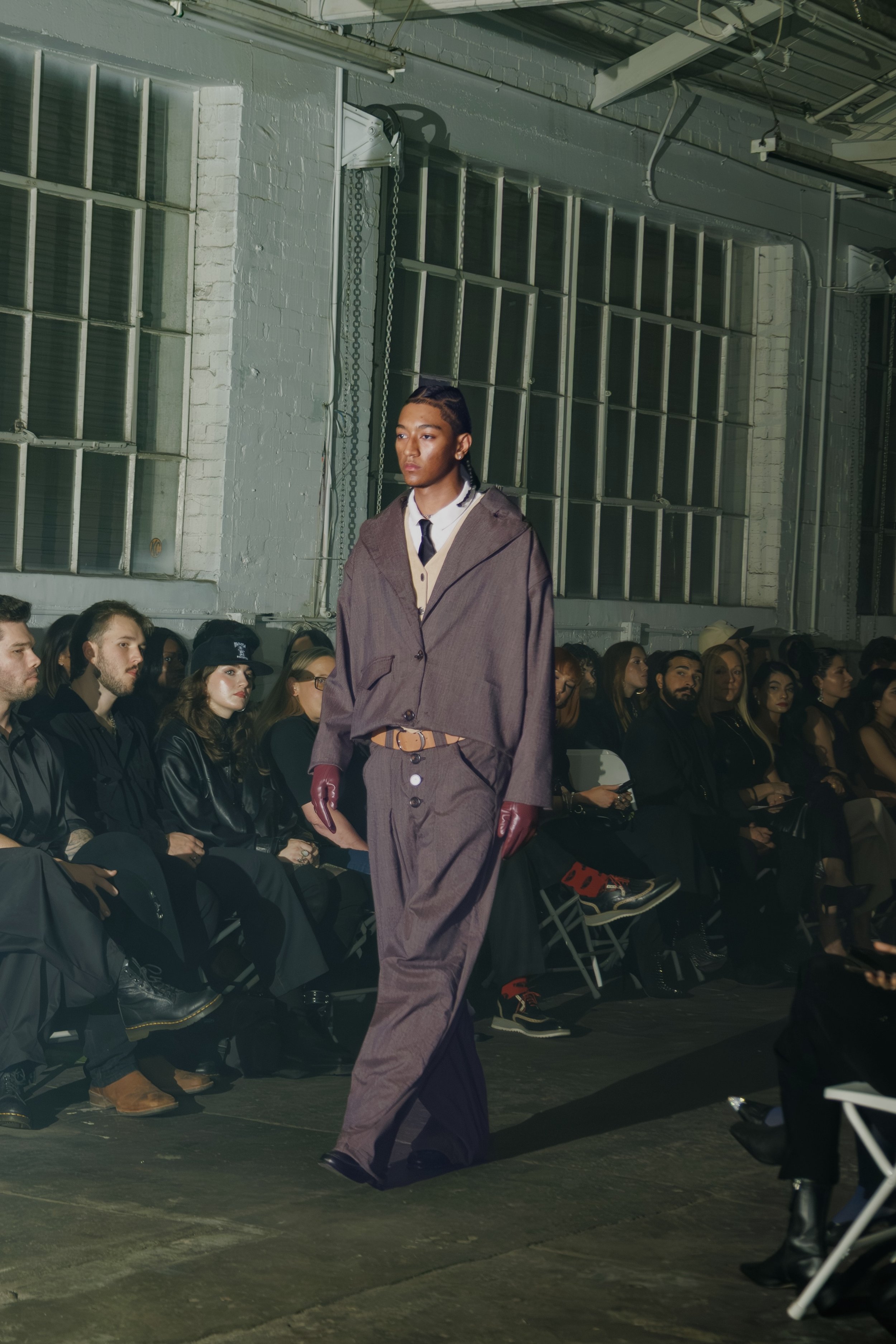 A male model walking on a runway during a fashion show, wearing a brown suit with wide-legged pants, a beige vest, a white shirt, and a black tie, with the audience seated on either side.