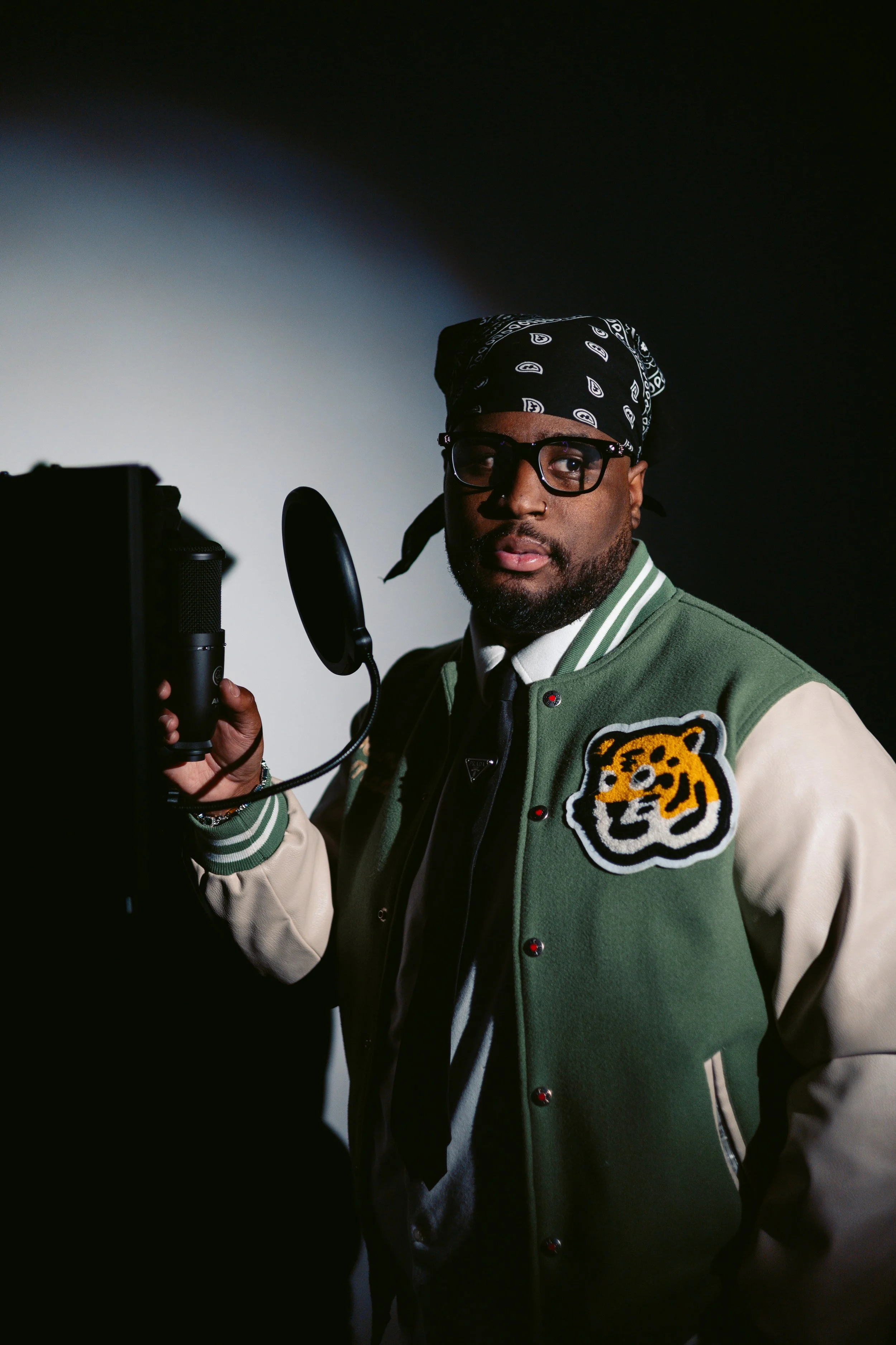 A man wearing glasses, a black bandana with white paisley pattern, and a green and cream varsity jacket with a tiger patch, standing in front of a microphone.