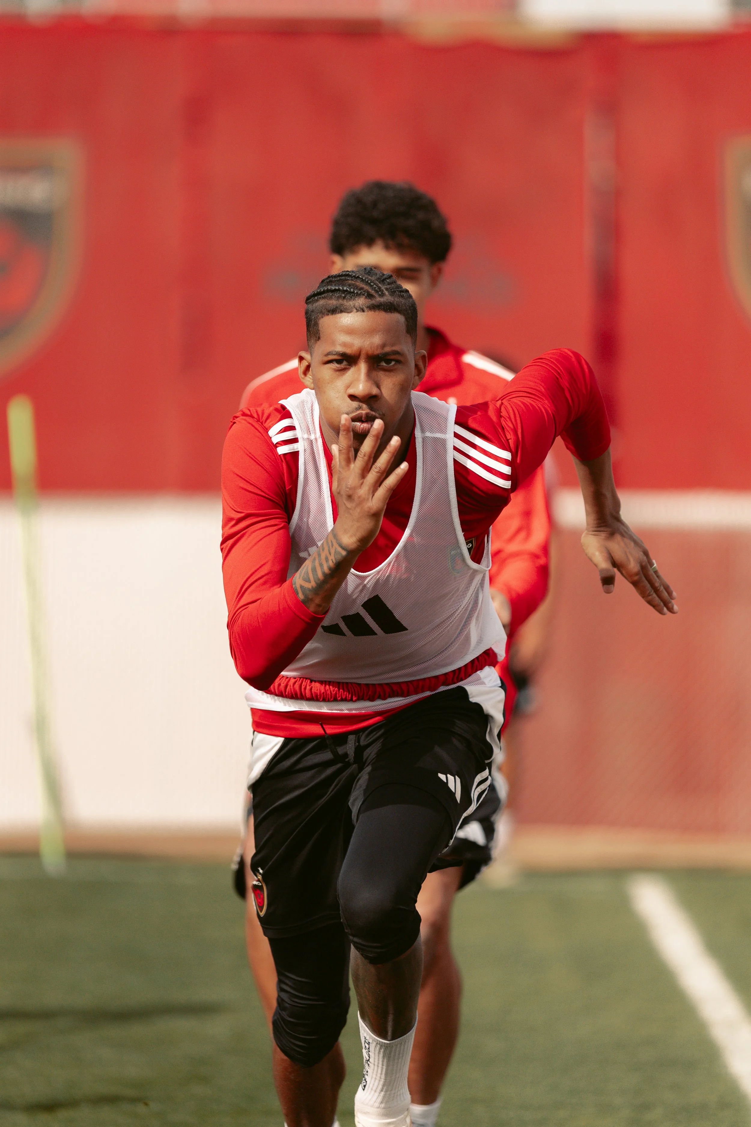 A group of football players in red jerseys practicing on a field, with the focus on a young man with braided hair and a white training vest looking forward as he runs.