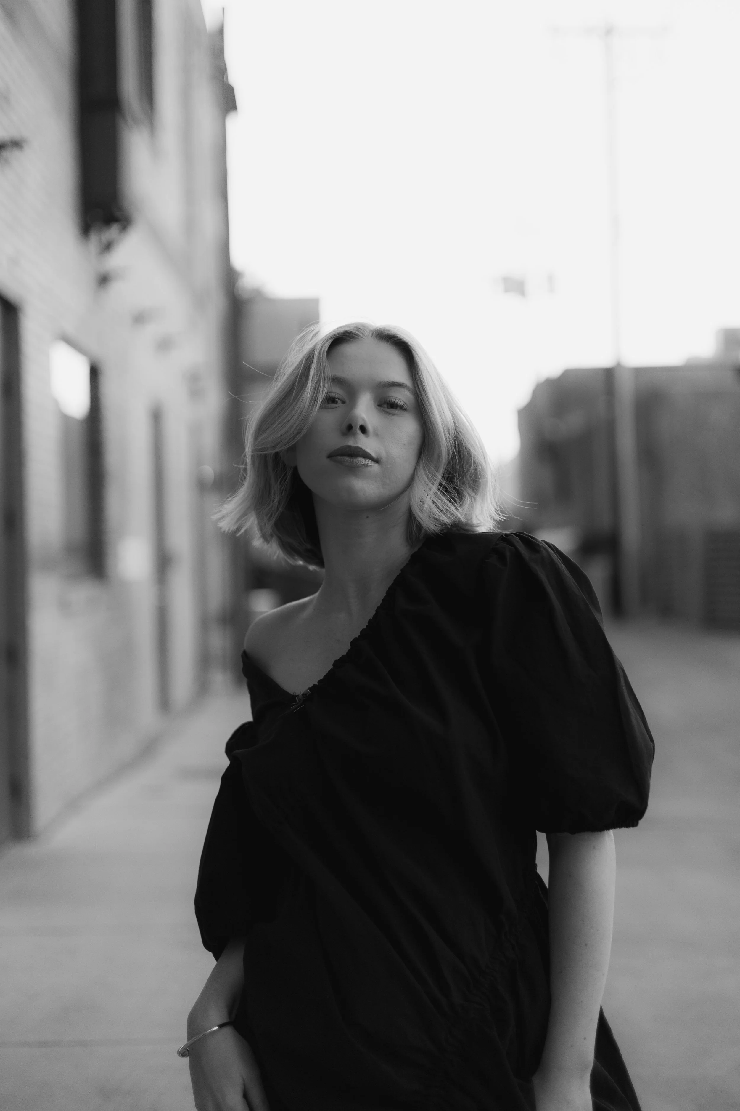 A black-and-white photograph of a young woman with shoulder-length hair, wearing a dark off-the-shoulder top, standing on a city street with the background showing blurred buildings and a sidewalk.