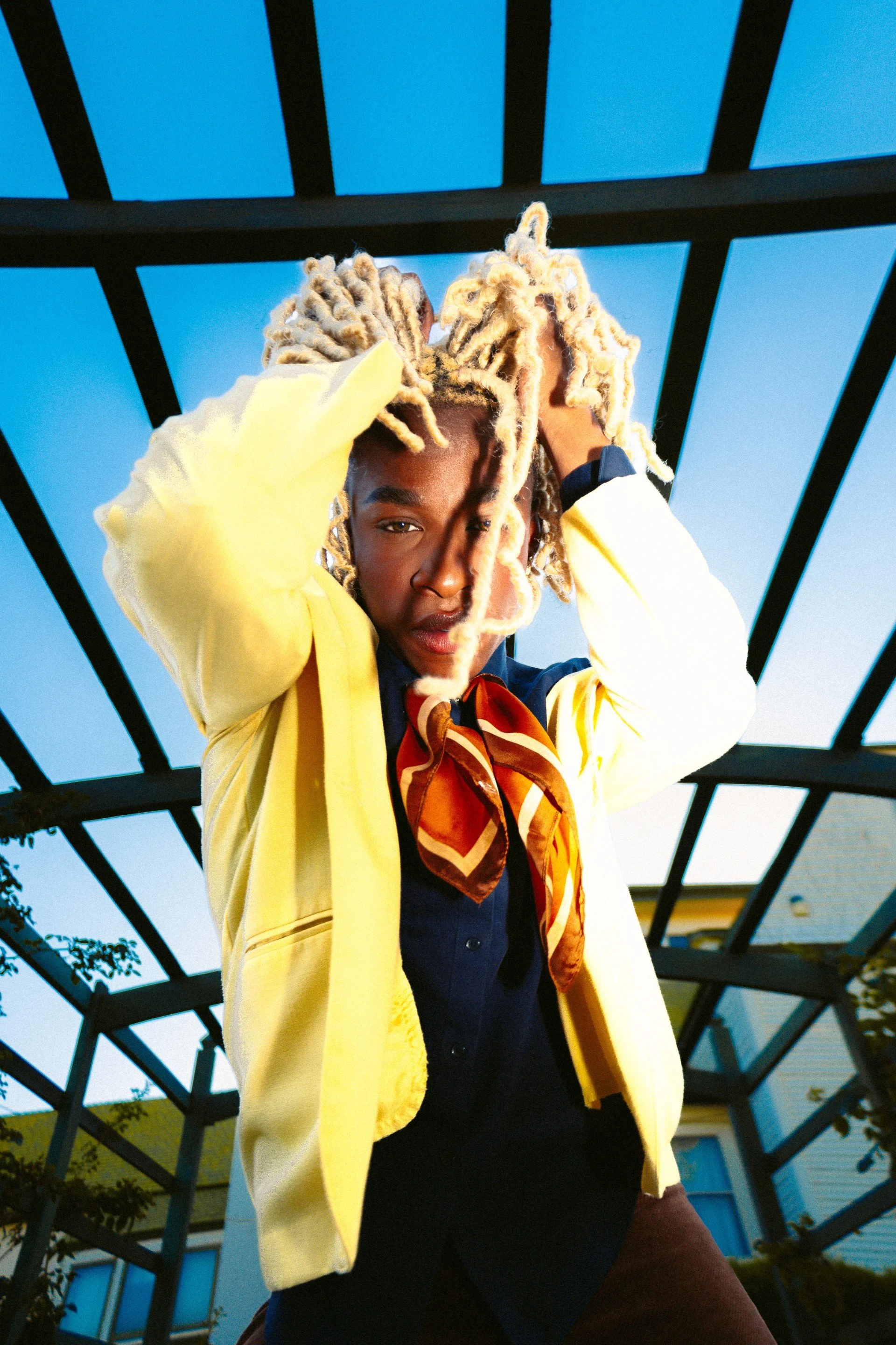 A young man in a blue blazer with gold cords, wearing a white shirt, standing on a porch with his arms resting on a red railing, looking thoughtfully to the side with a brick wall and green door behind him.