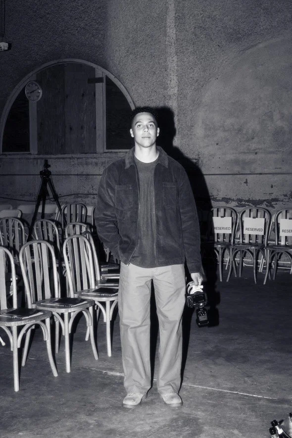A man standing in a room with chairs, holding a camera in his right hand, and looking at the camera. The room has a large arch window and a clock.