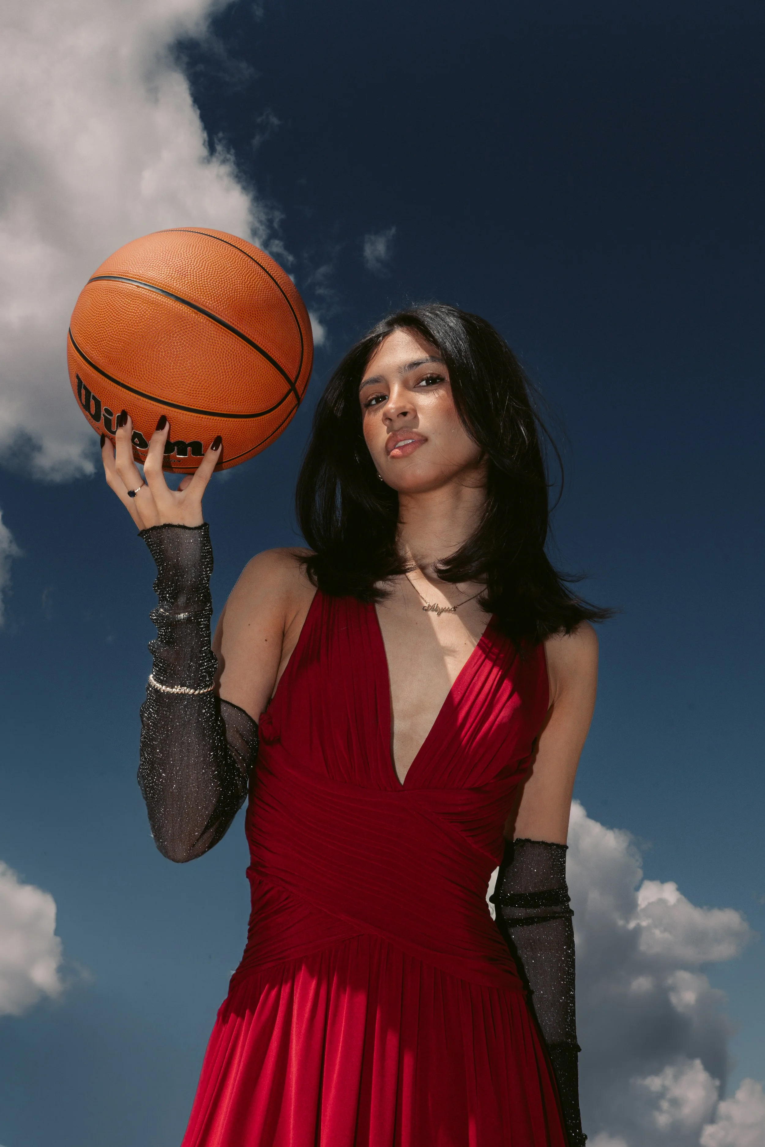 A woman in a red dress holding a basketball against a partly cloudy sky.