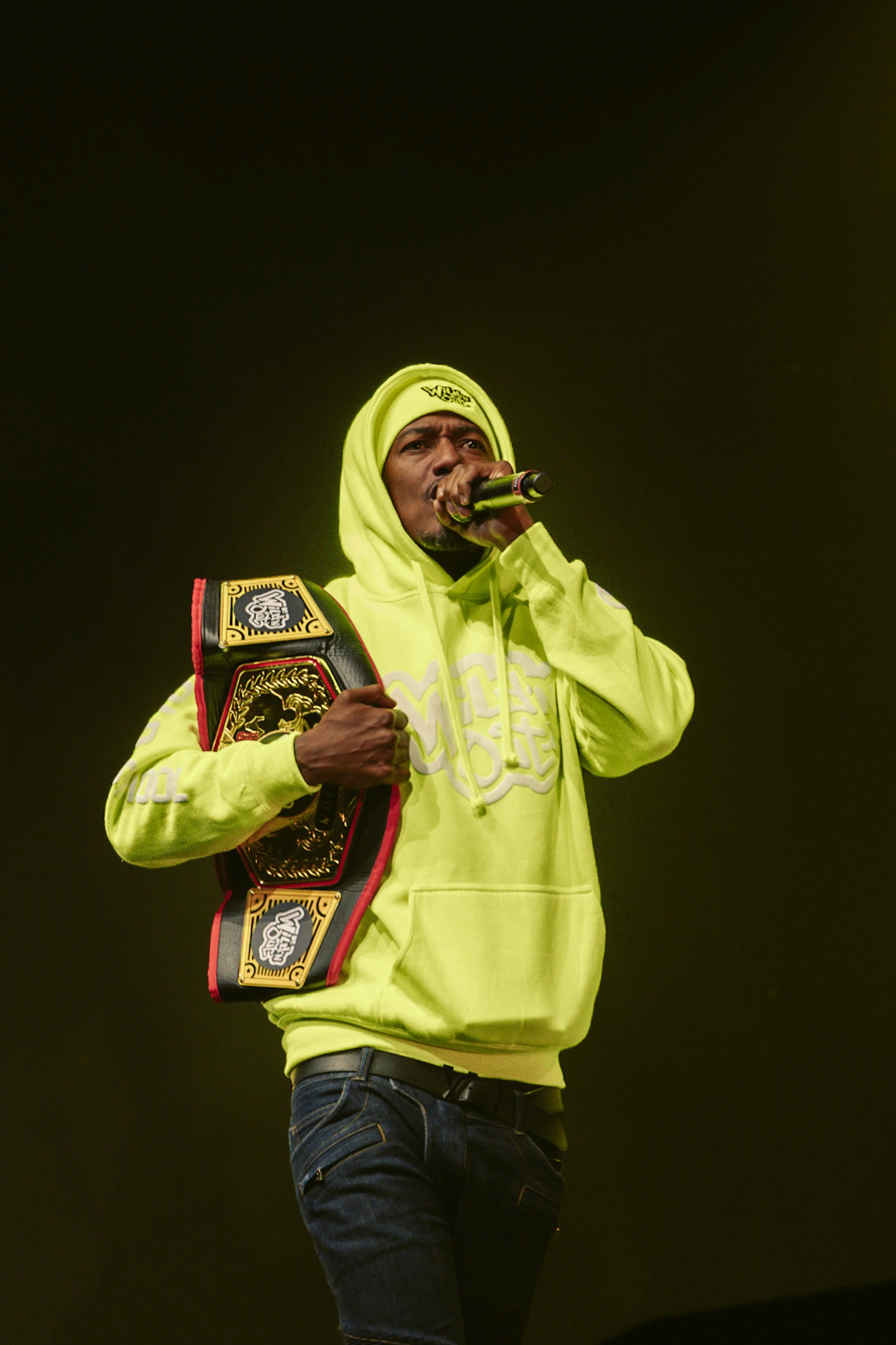 A man wearing a yellow hoodie and a neon yellow beanie holds a championship belt and speaks into a microphone on stage.