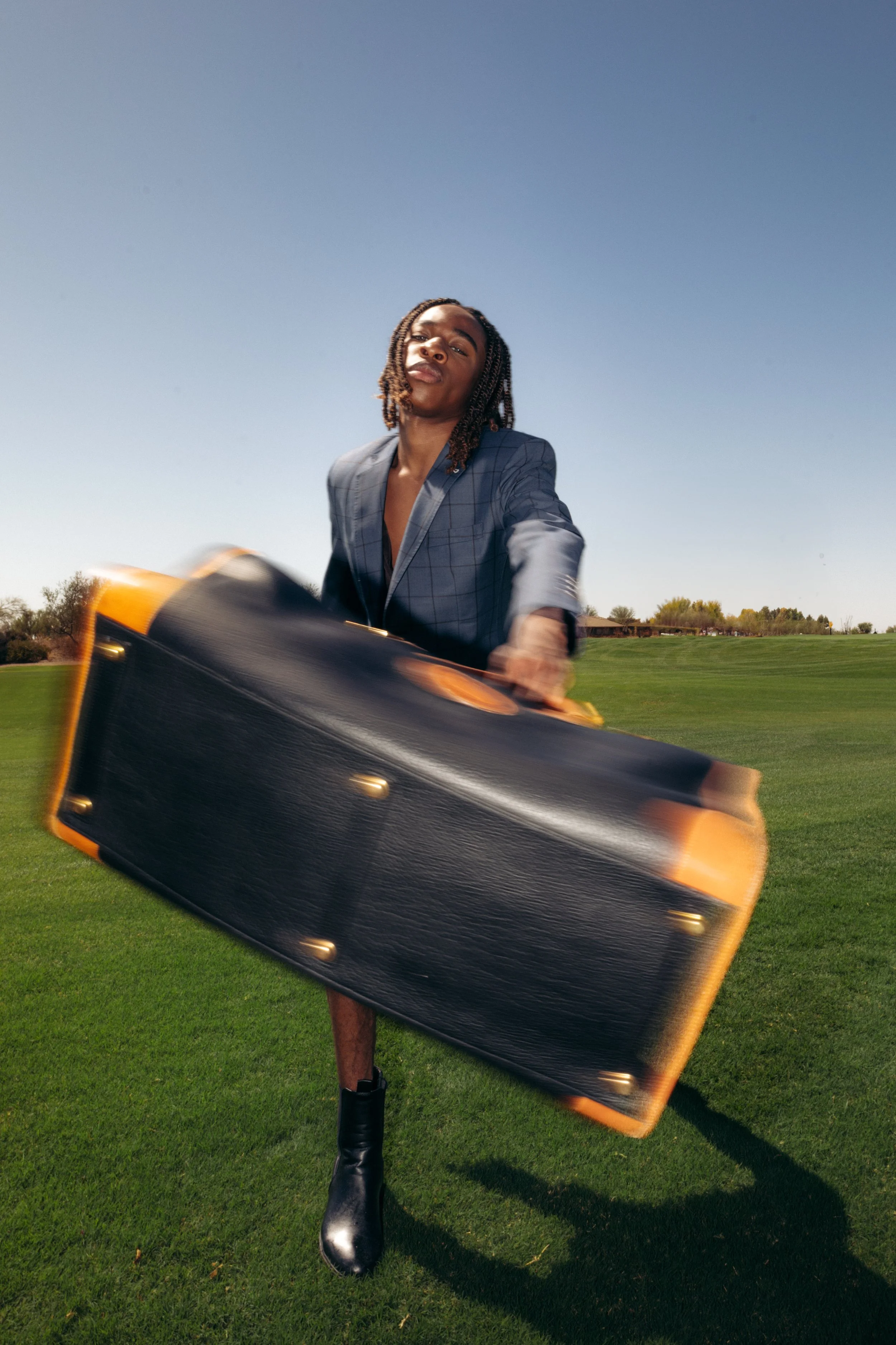 A person with long braids, wearing a blue checkered blazer and shiny black boots, standing on green grass under a clear blue sky, holding a large briefcase that is blurred due to movement.
