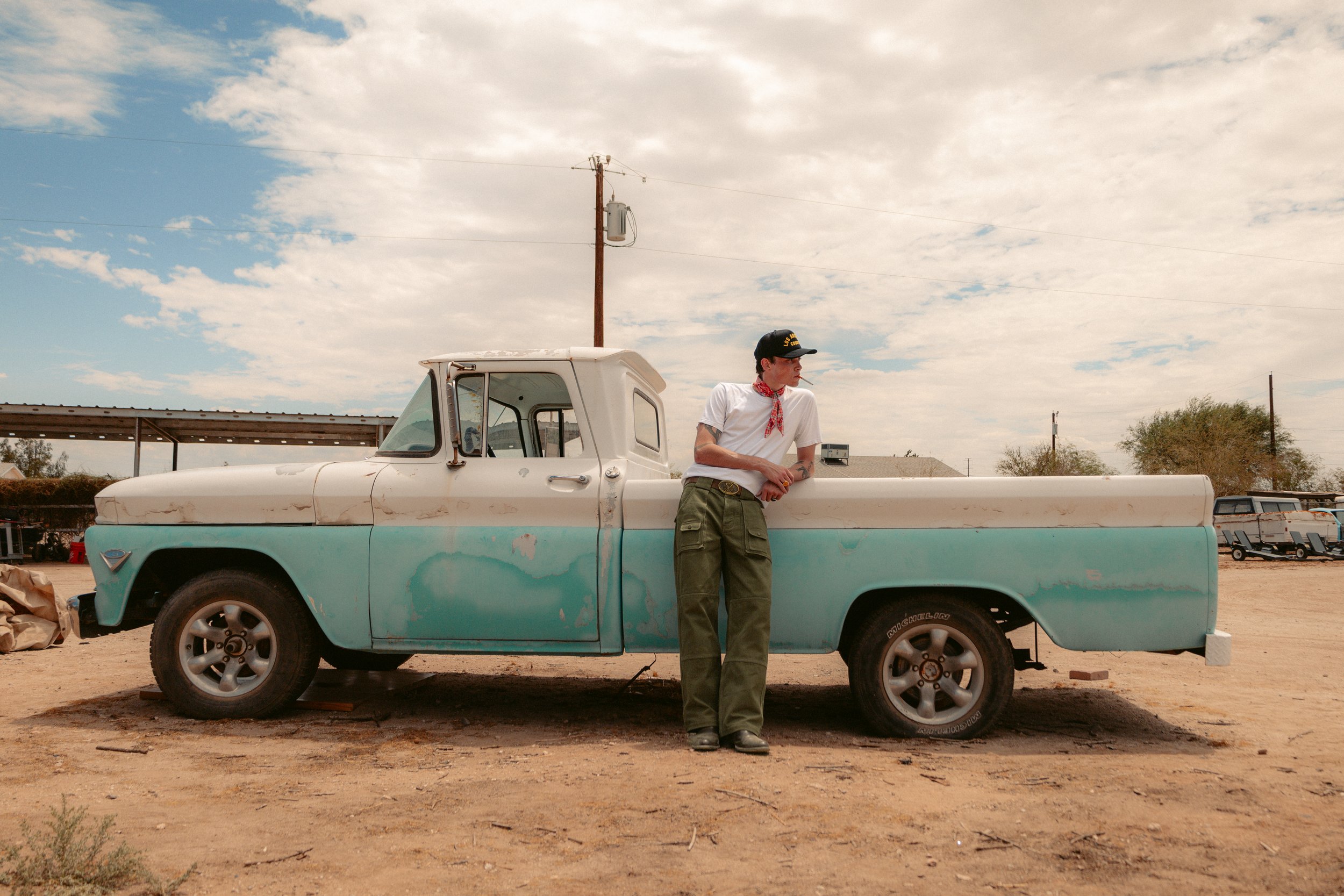 A young man wearing a black cap, white t-shirt, red bandana, and green pants leaning against a vintage pickup truck in a rural area with a partly cloudy sky overhead.