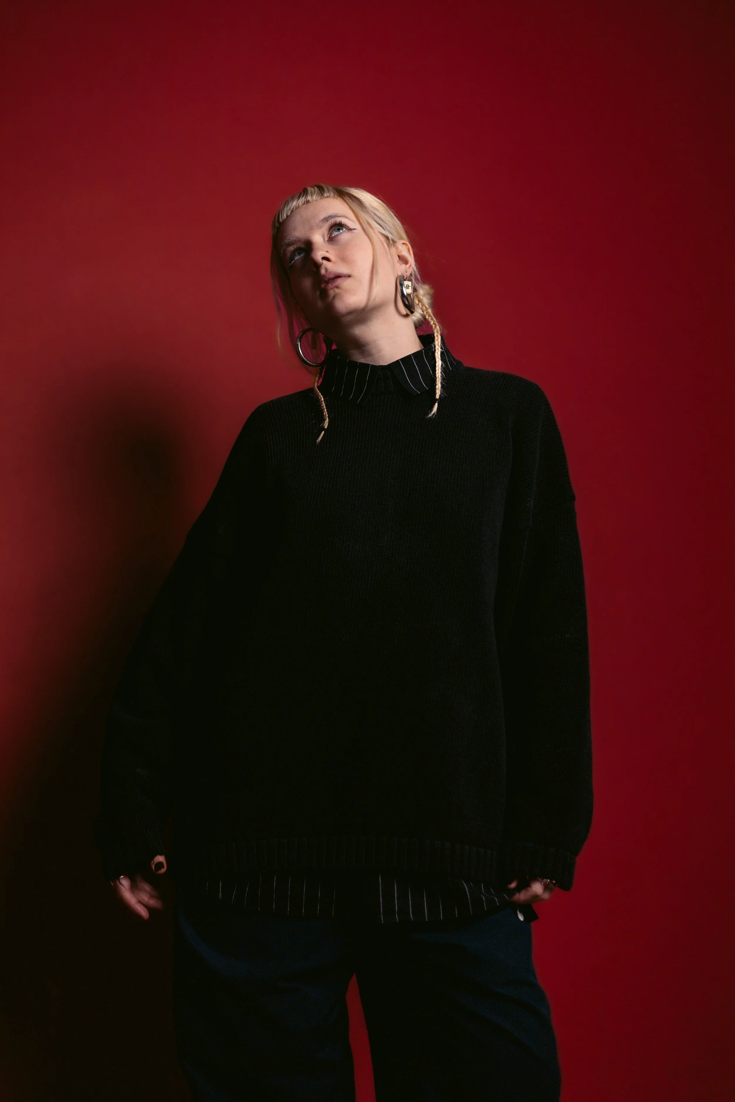 A woman with blonde hair and earrings, wearing a black sweater, standing against a red background.