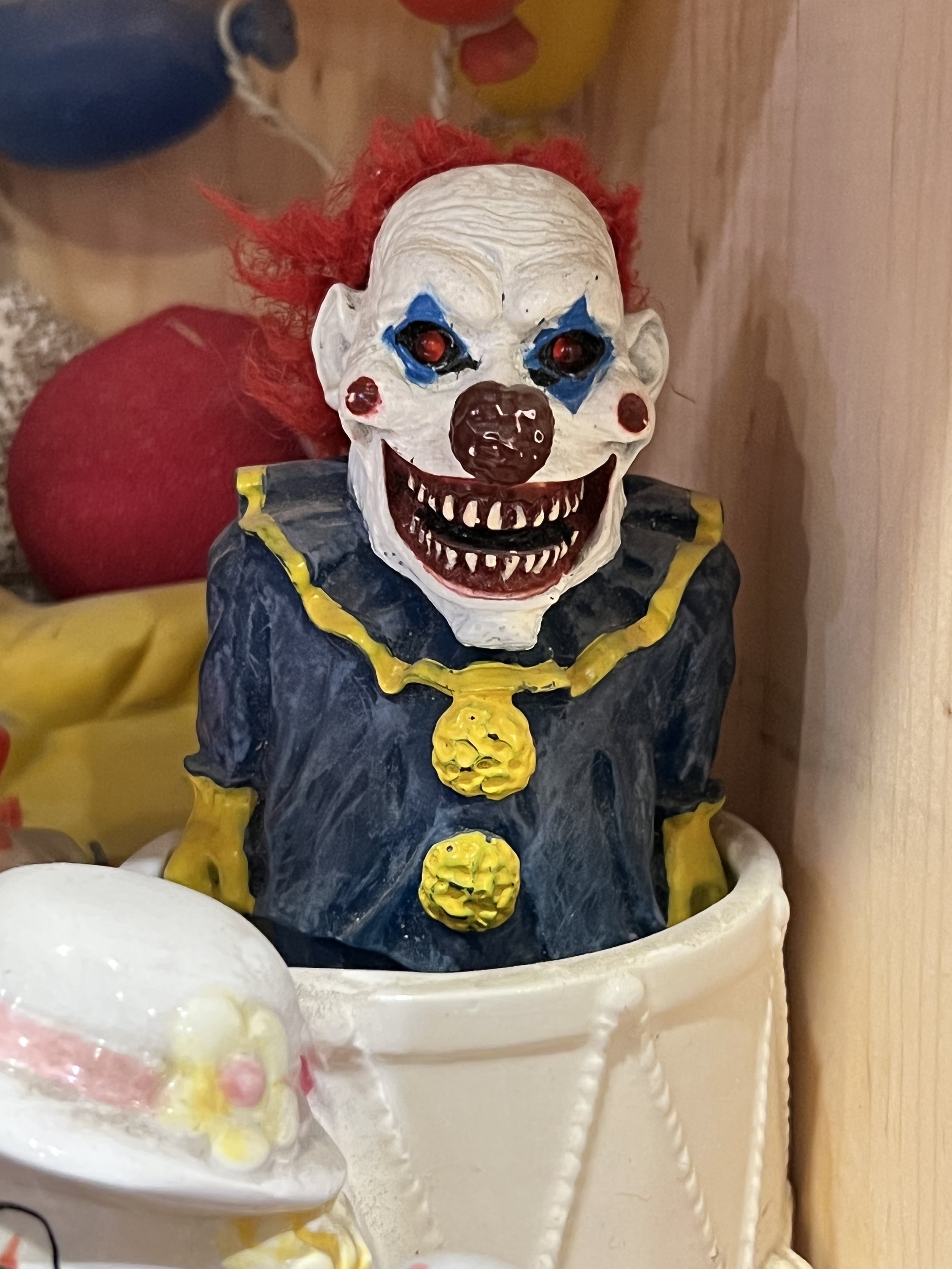 A clown-themed figurine with a creepy clown face, red hair, and clown costume, sitting inside a white container.