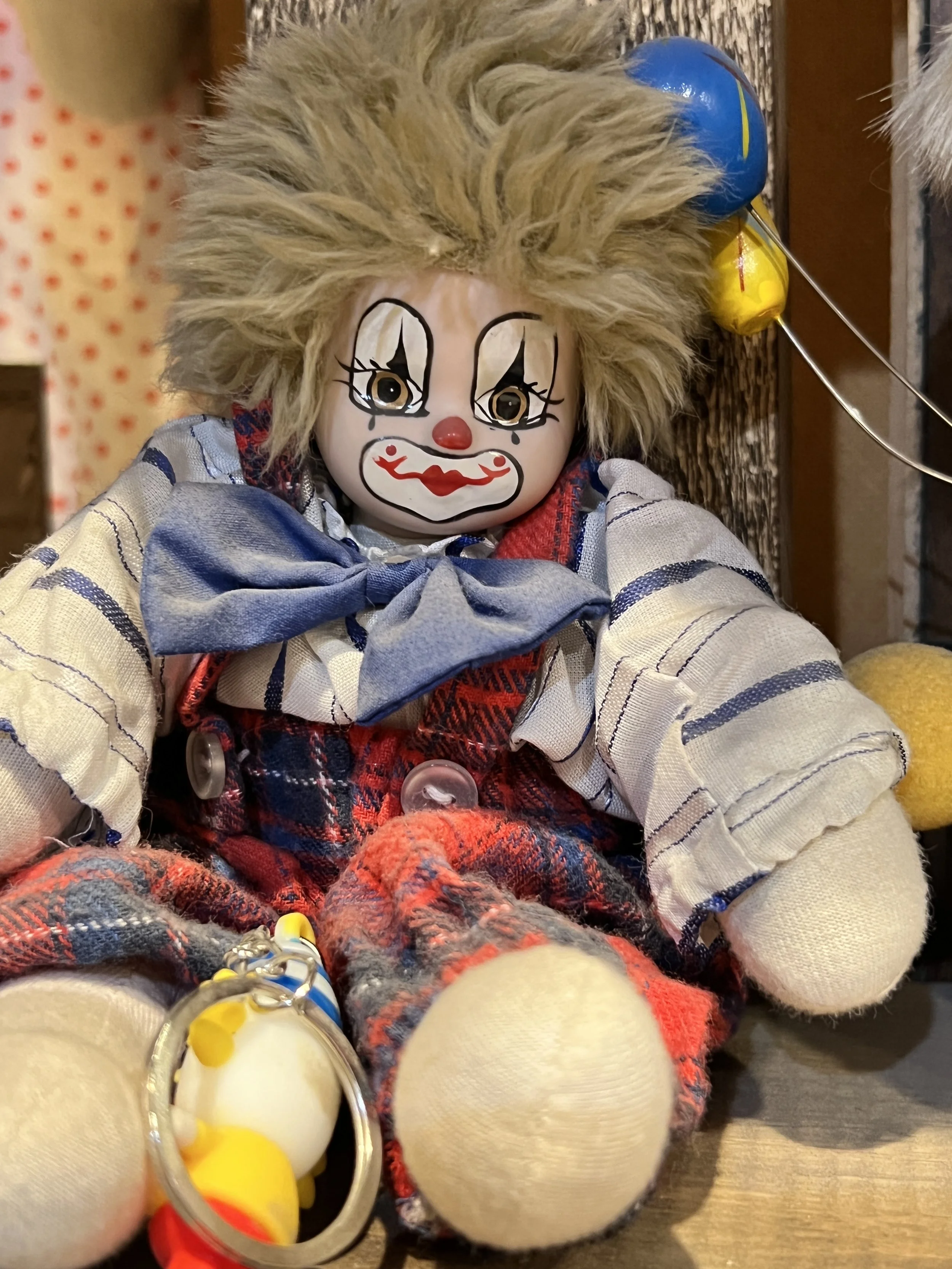 A clown doll with messy blond hair, painted face, big red lips, and exaggerated eyes. It is dressed in a striped shirt, plaid pants, and a blue bow tie, sitting among other objects.