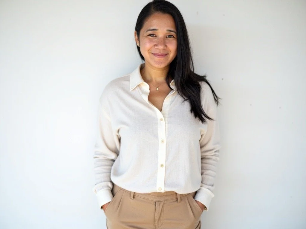 A woman with long black hair, dressed in a cream-colored button-down shirt and beige pants, standing against a plain white wall, smiling with her hands in her pockets.