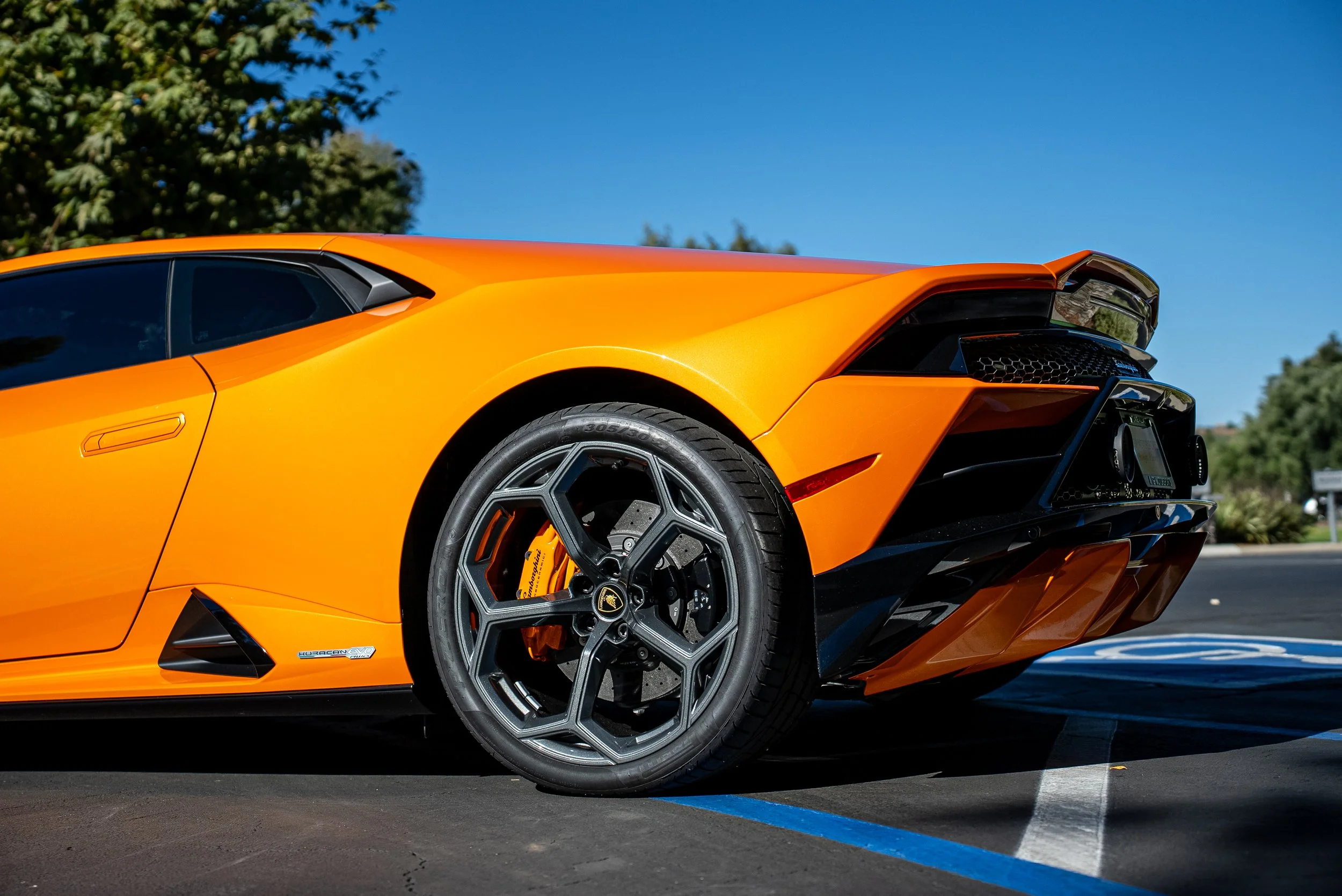 Orange Lamborghini sports car parked on the road with trees and blue sky in the background.
