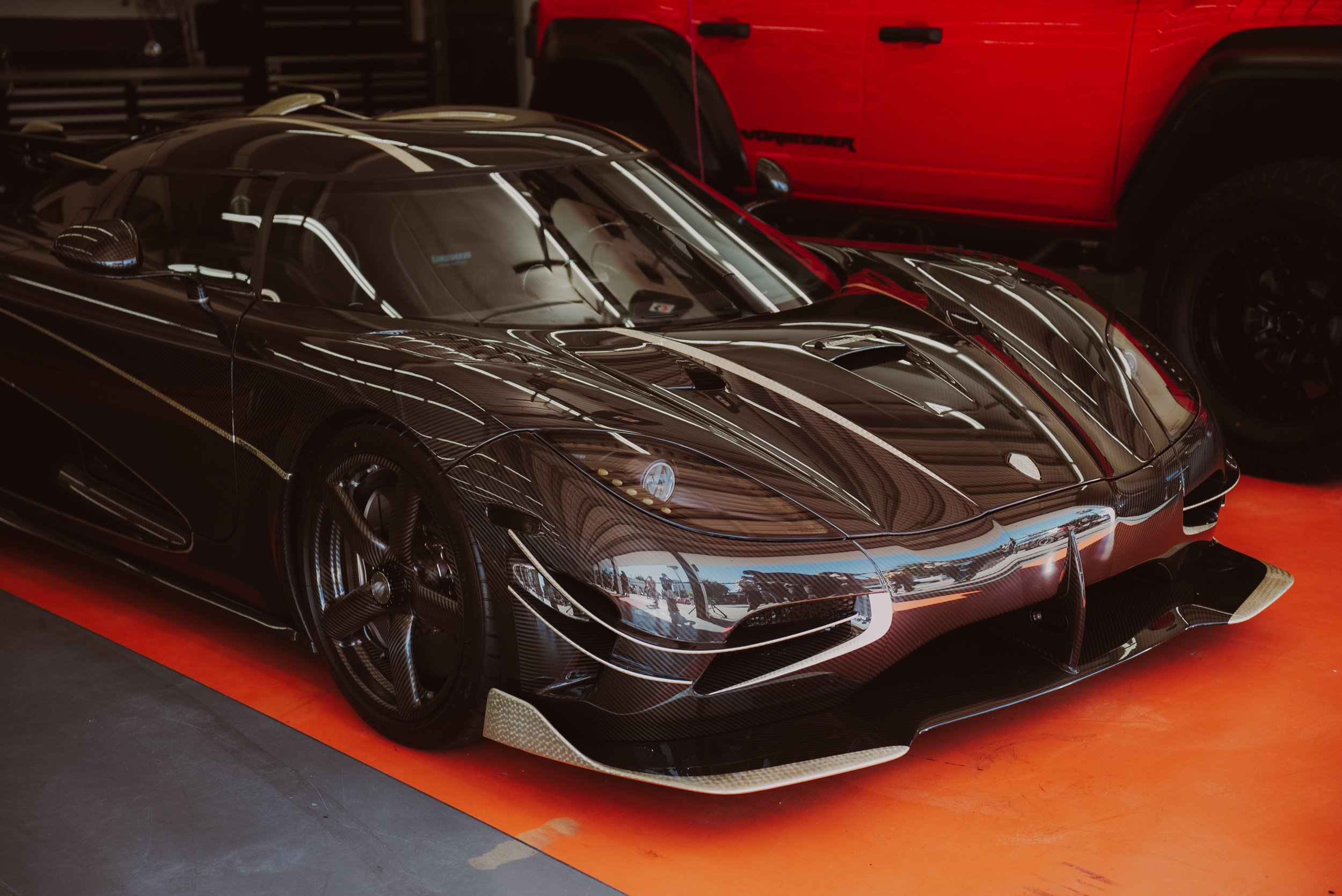 A sleek black racing car with aerodynamic features and carbon fiber accents, parked indoors on an orange surface, with a red vehicle partially visible behind it.