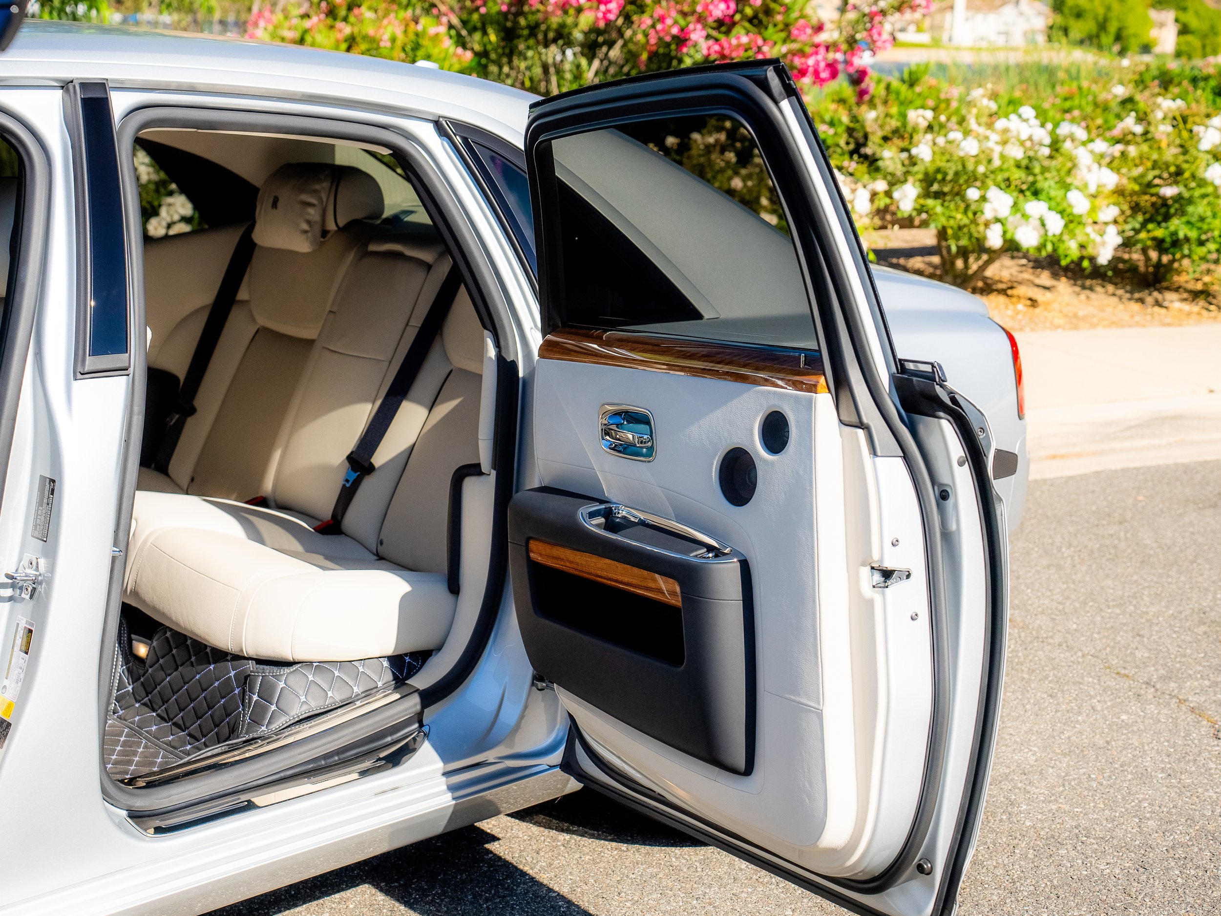 Open door of a luxury car showing beige leather interior and decorative wood paneling with outdoor garden and pink flowers in the background.