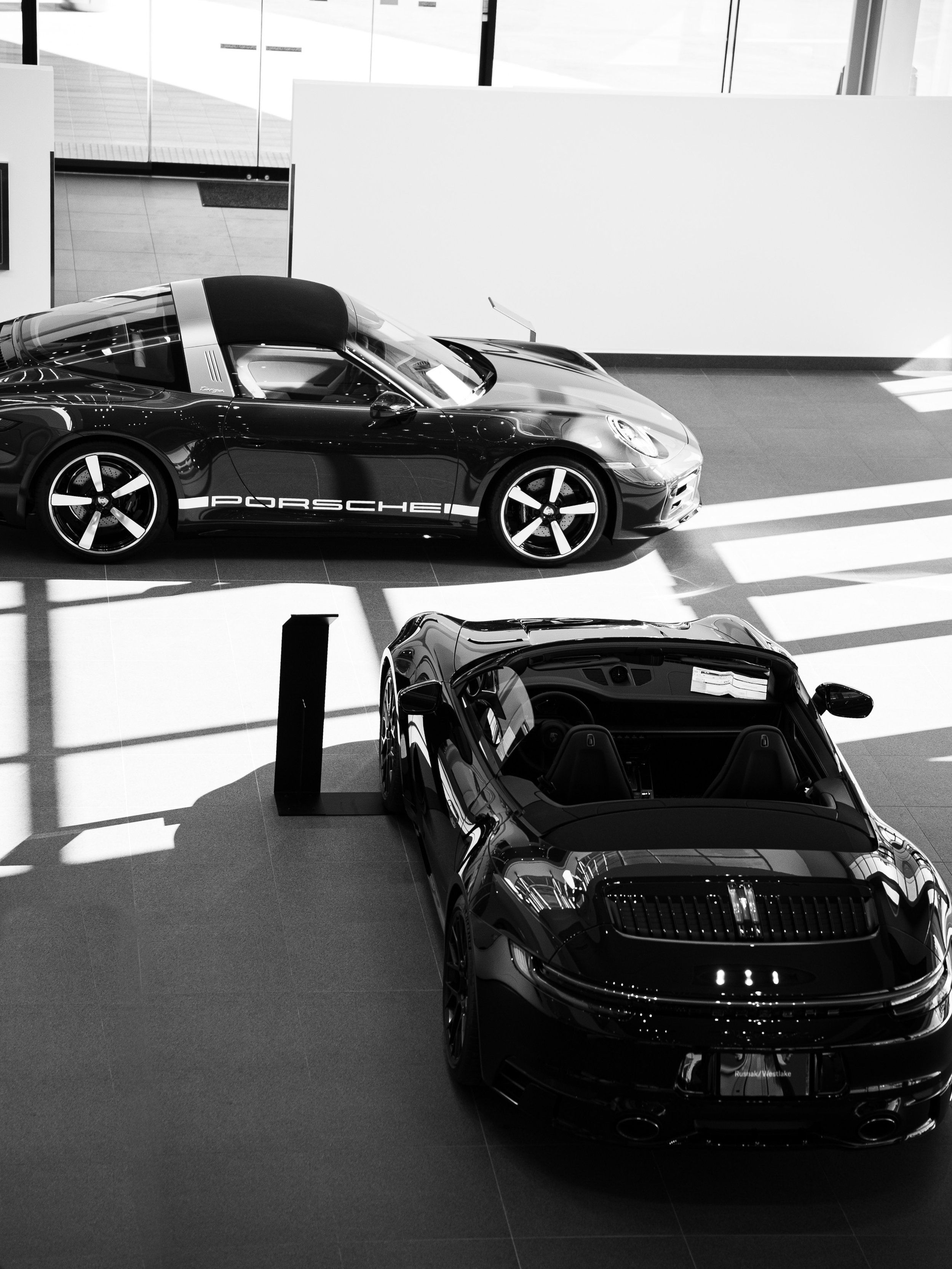 A black Porsche sports car with a convertible top on display in a showroom, with another right behind it and sunlight casting shadows on the floor.