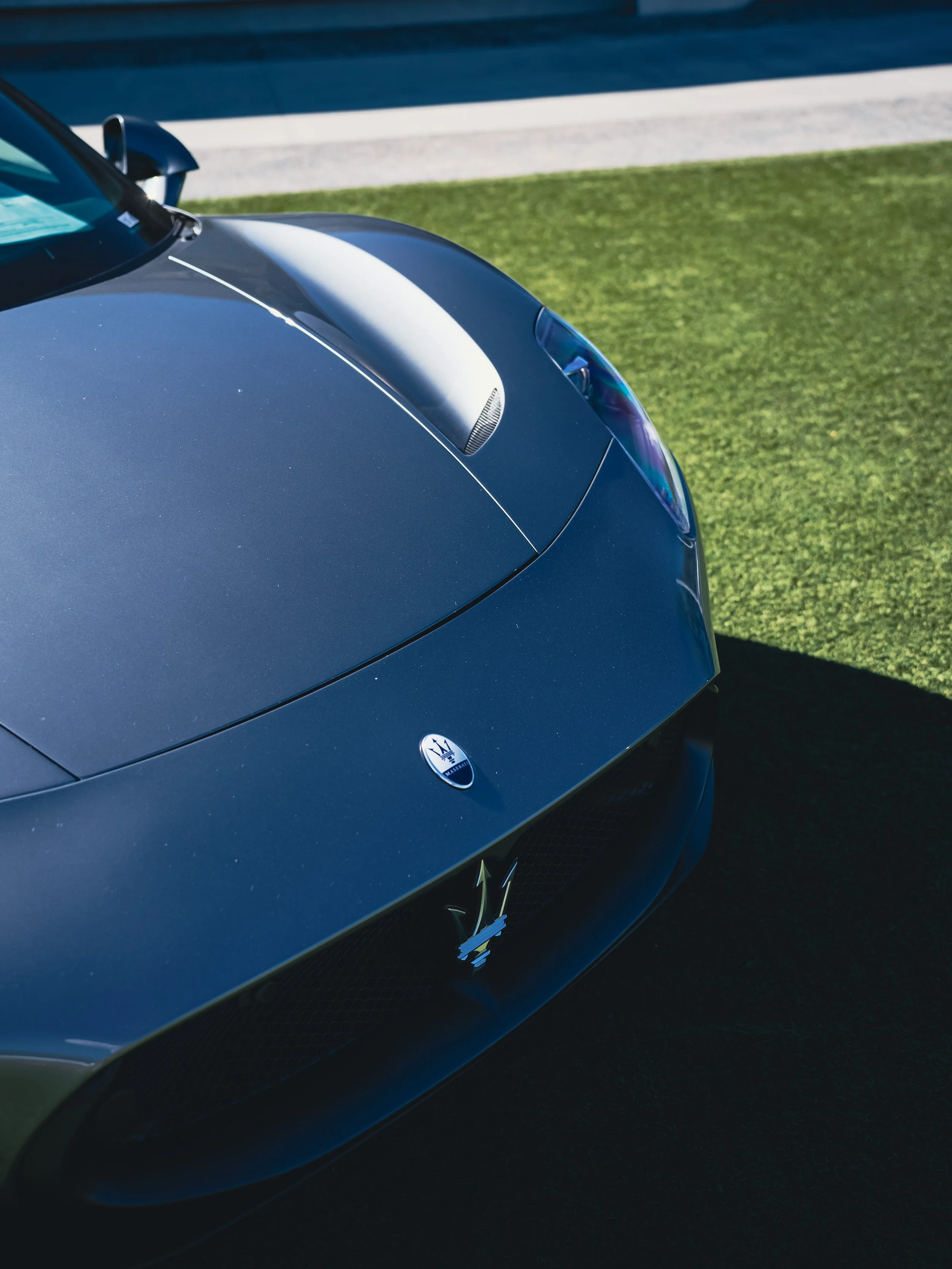 Close-up of a dark blue Maserati sports car on grass near a sidewalk, showing the front hood, headlight, and Maserati emblem.