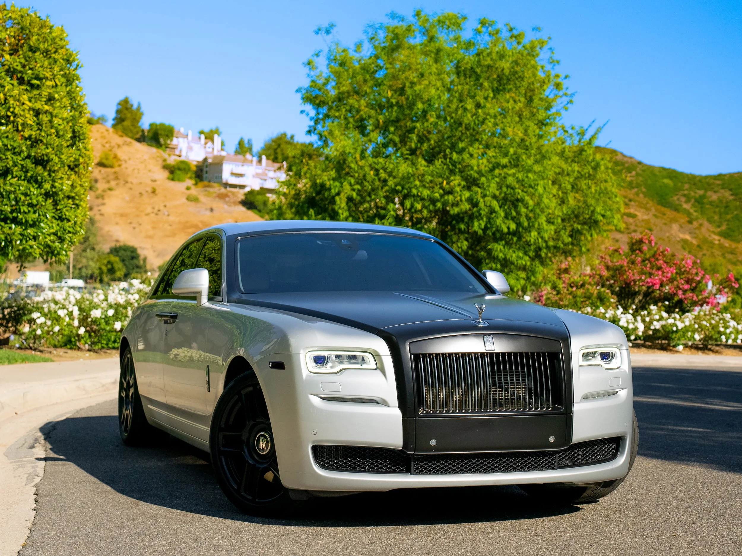 Luxury silver Rolls-Royce car parked on a street with greenery and scenic hillside in the background.