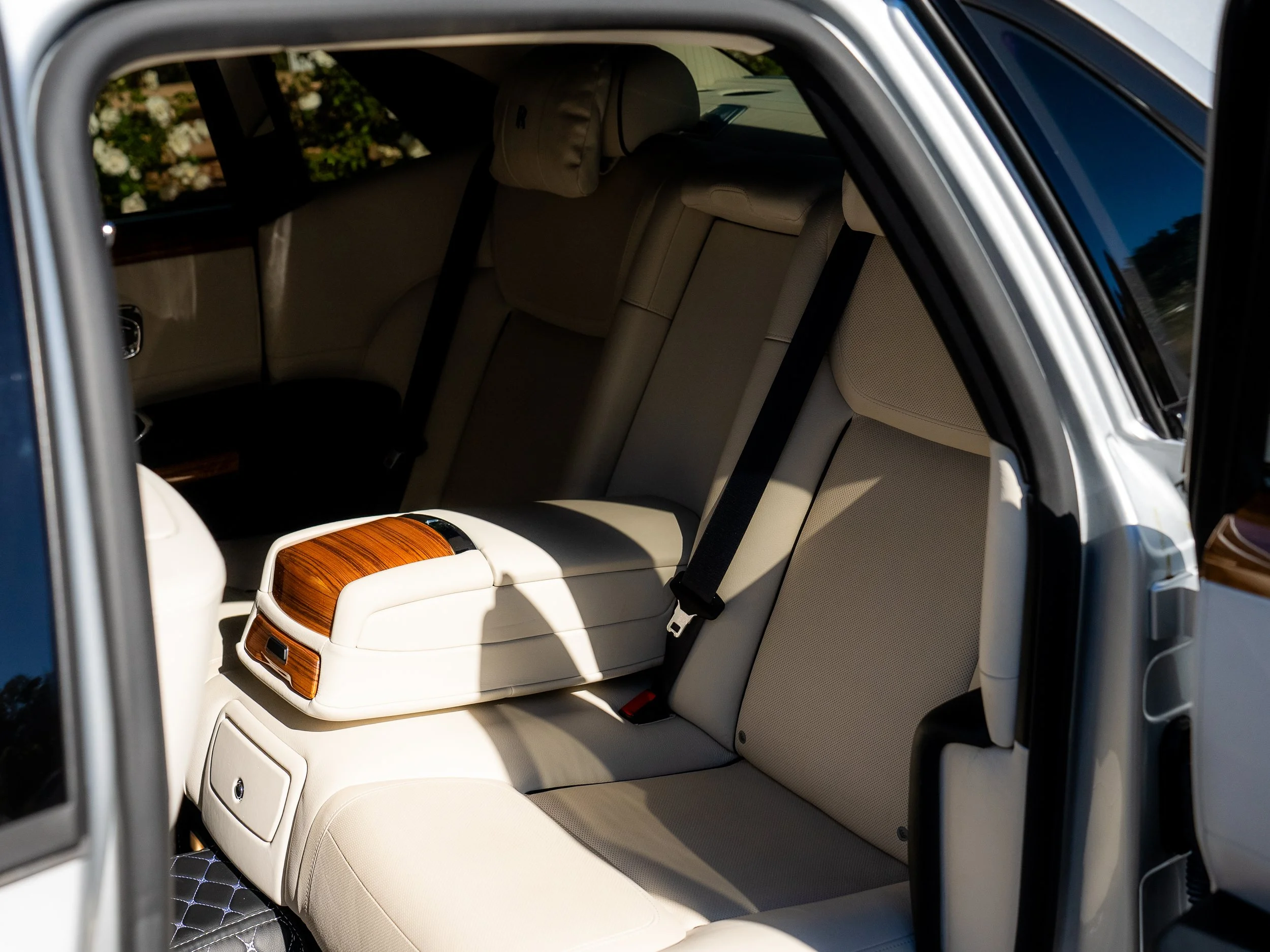 Interior of a luxury car's back seat with beige leather seats and wooden accents, visible through an open car door.
