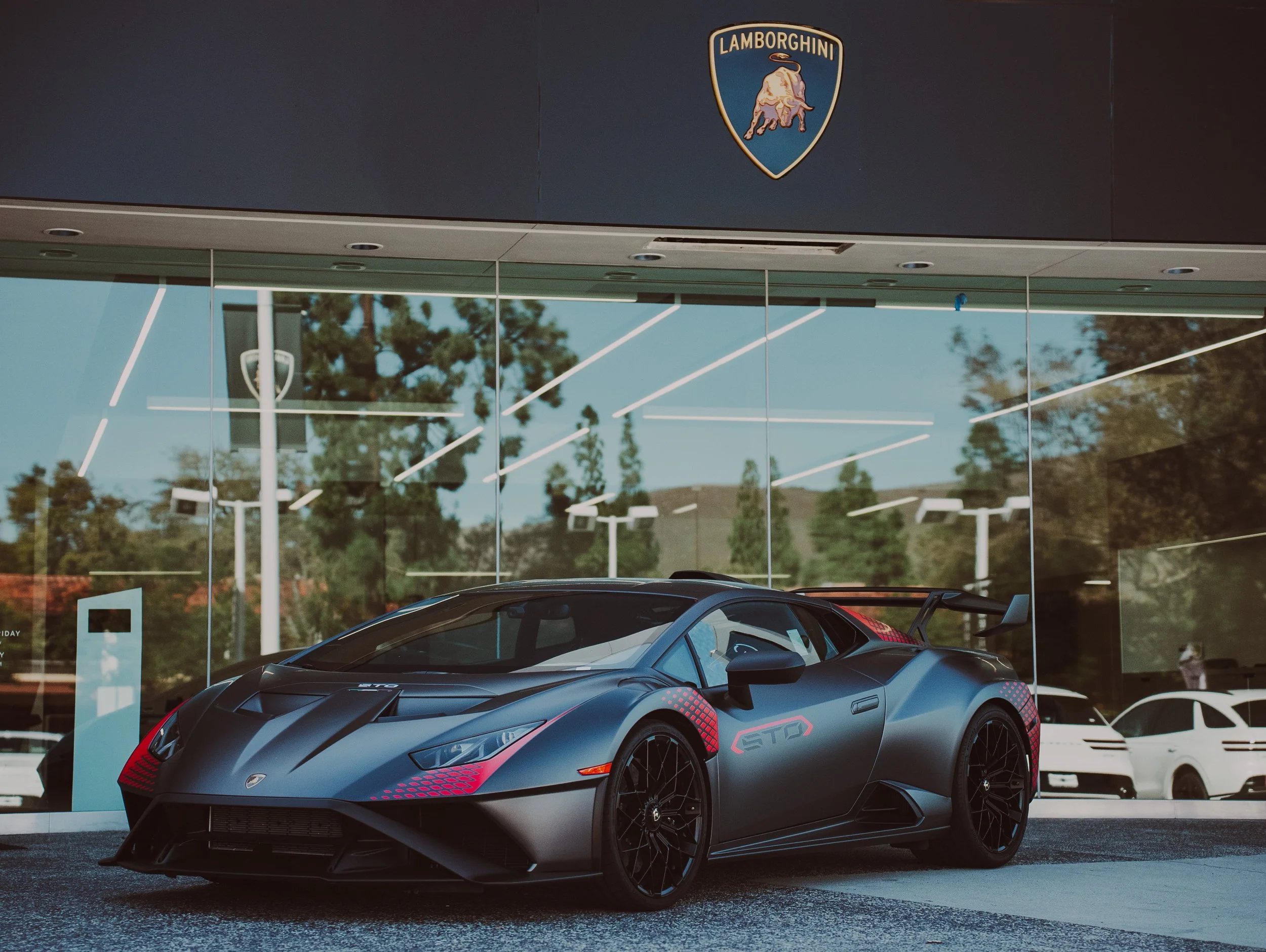 A matte black Lamborghini sports car parked in front of a dealership with the Lamborghini logo displayed above.