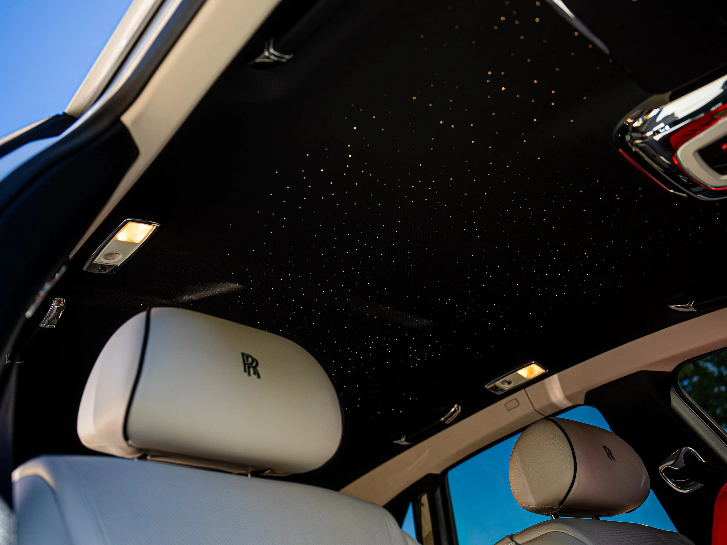 The interior of a luxury car featuring a black ceiling with tiny LED lights resembling stars, beige seats with the Rolls-Royce logo, and a panoramic window showing a blue sky outside.