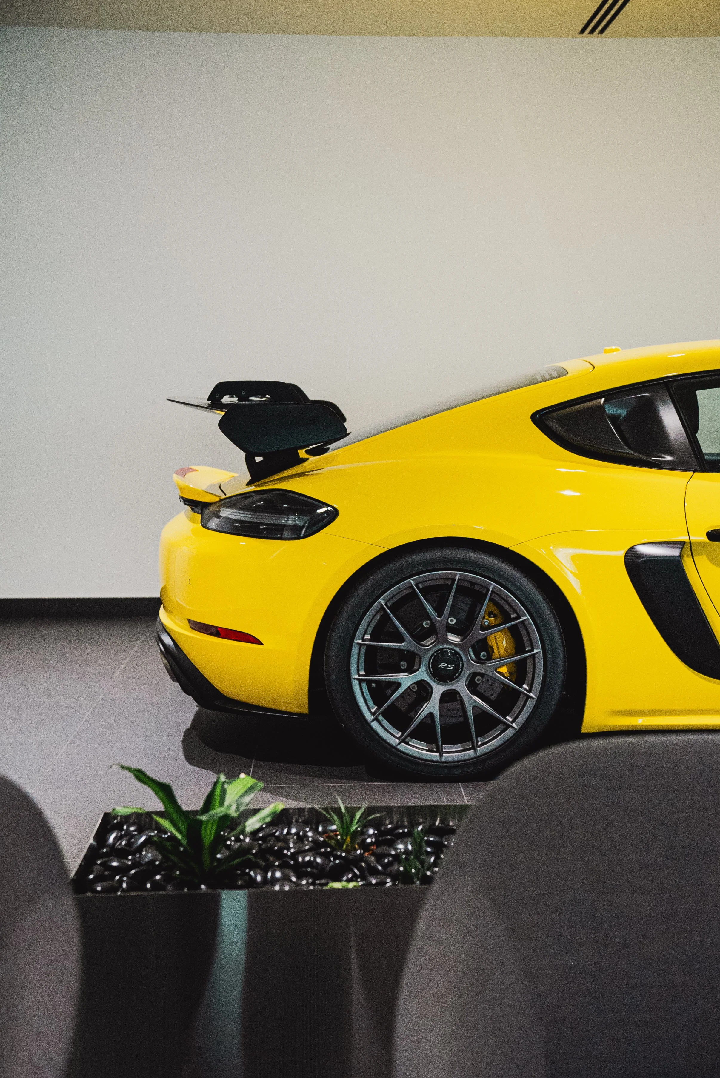 Close-up of a bright yellow sports car in an indoor showroom, showing the rear side with a large rear wing, black side vents, and intricate gray alloy wheels.