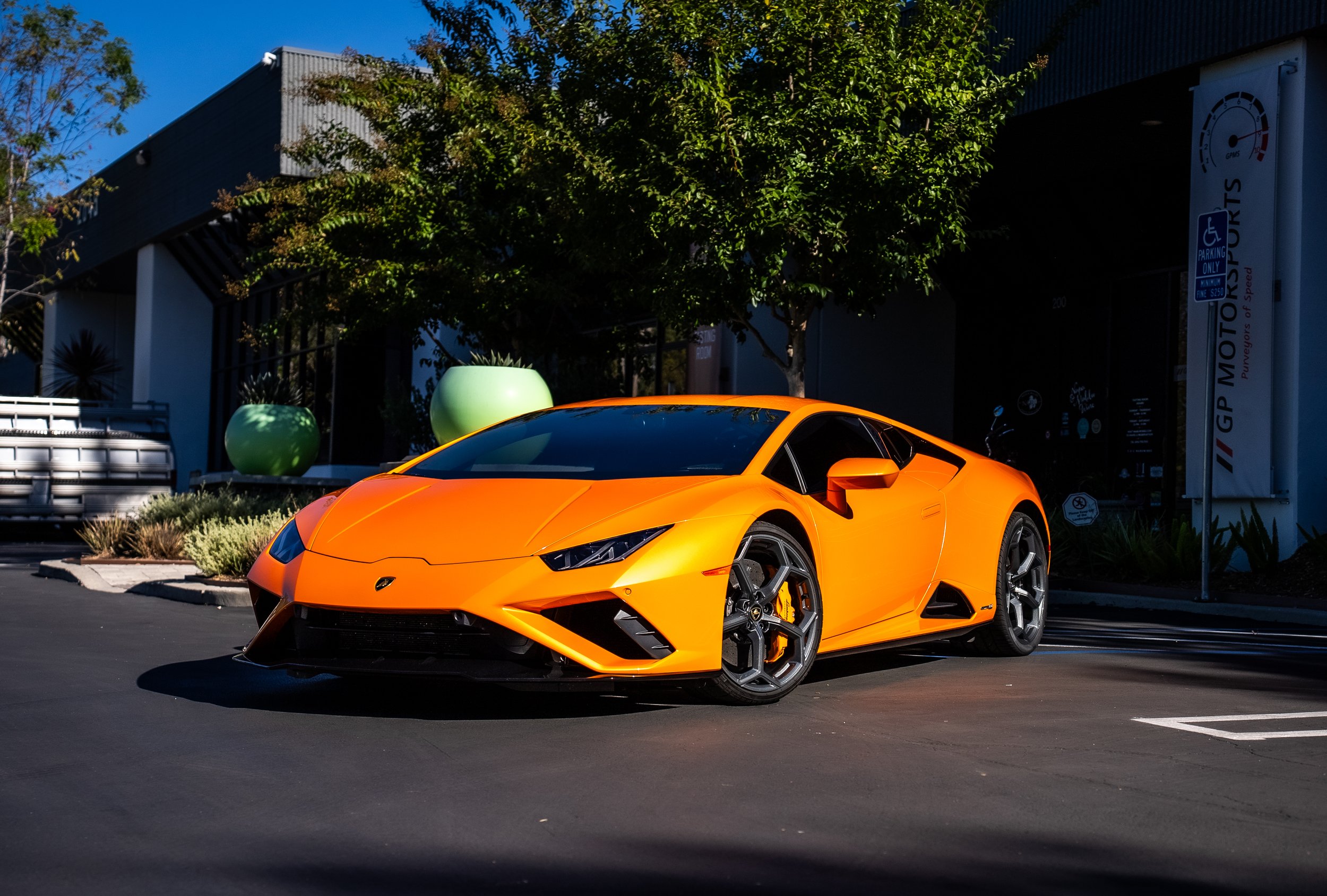Orange Lamborghini sports car parked in an outdoor lot near a building with green decorative plants.