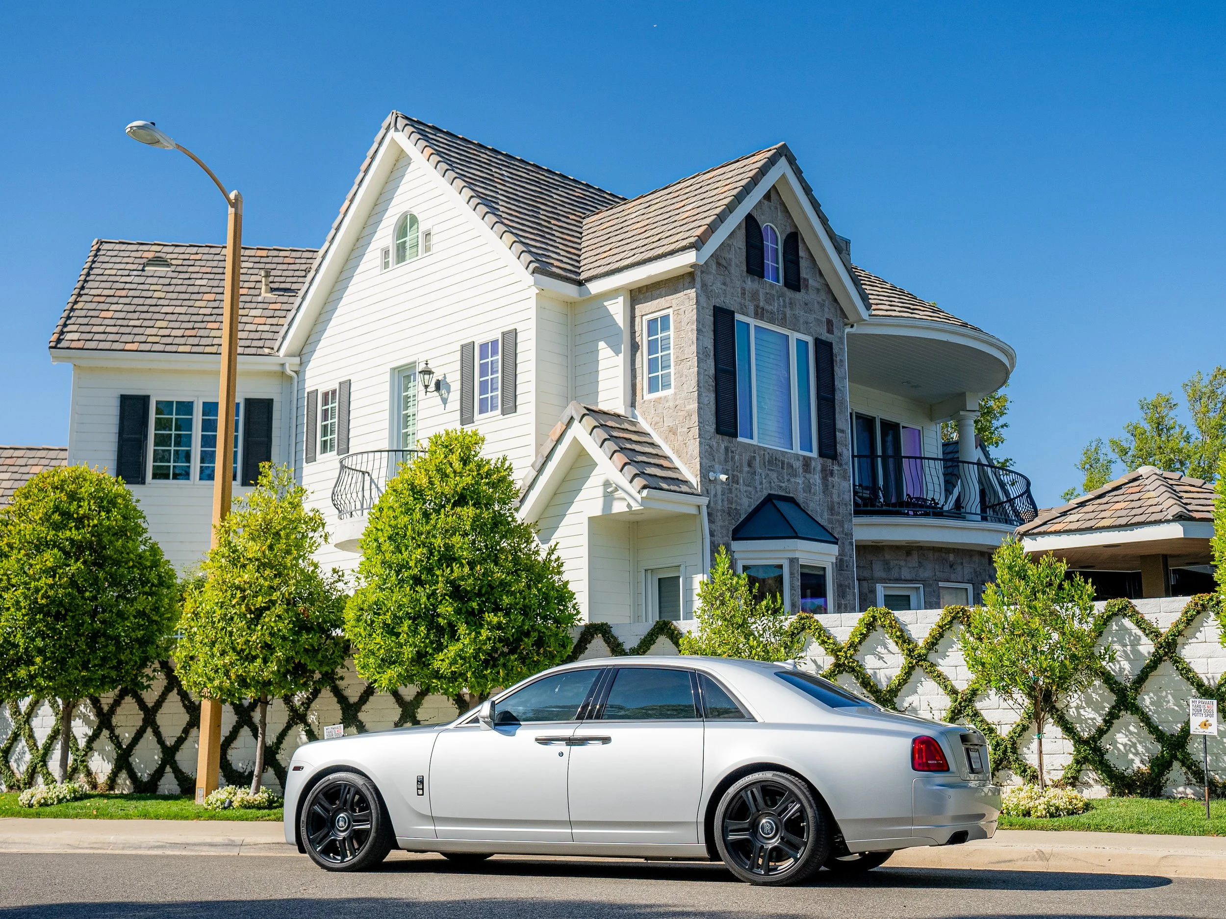 A silver luxury car parked on the street in front of a large multi-story house with beige siding, black shutters, a stone facade, and a tiled roof, with well-maintained greenery and trees surrounding the property.