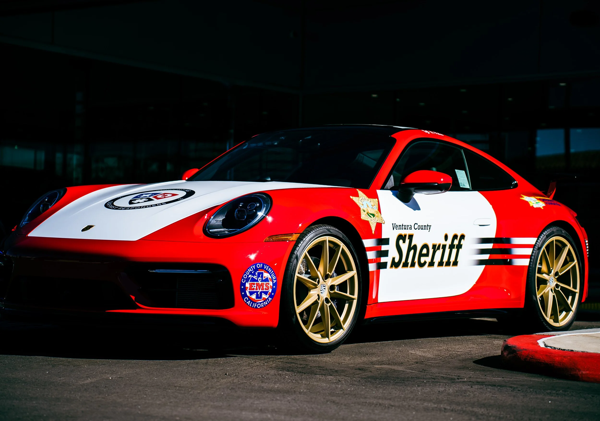 Red and white Porsche sports car with Ventura County Sheriff's Department markings, gold wheels, and emergency service badges.