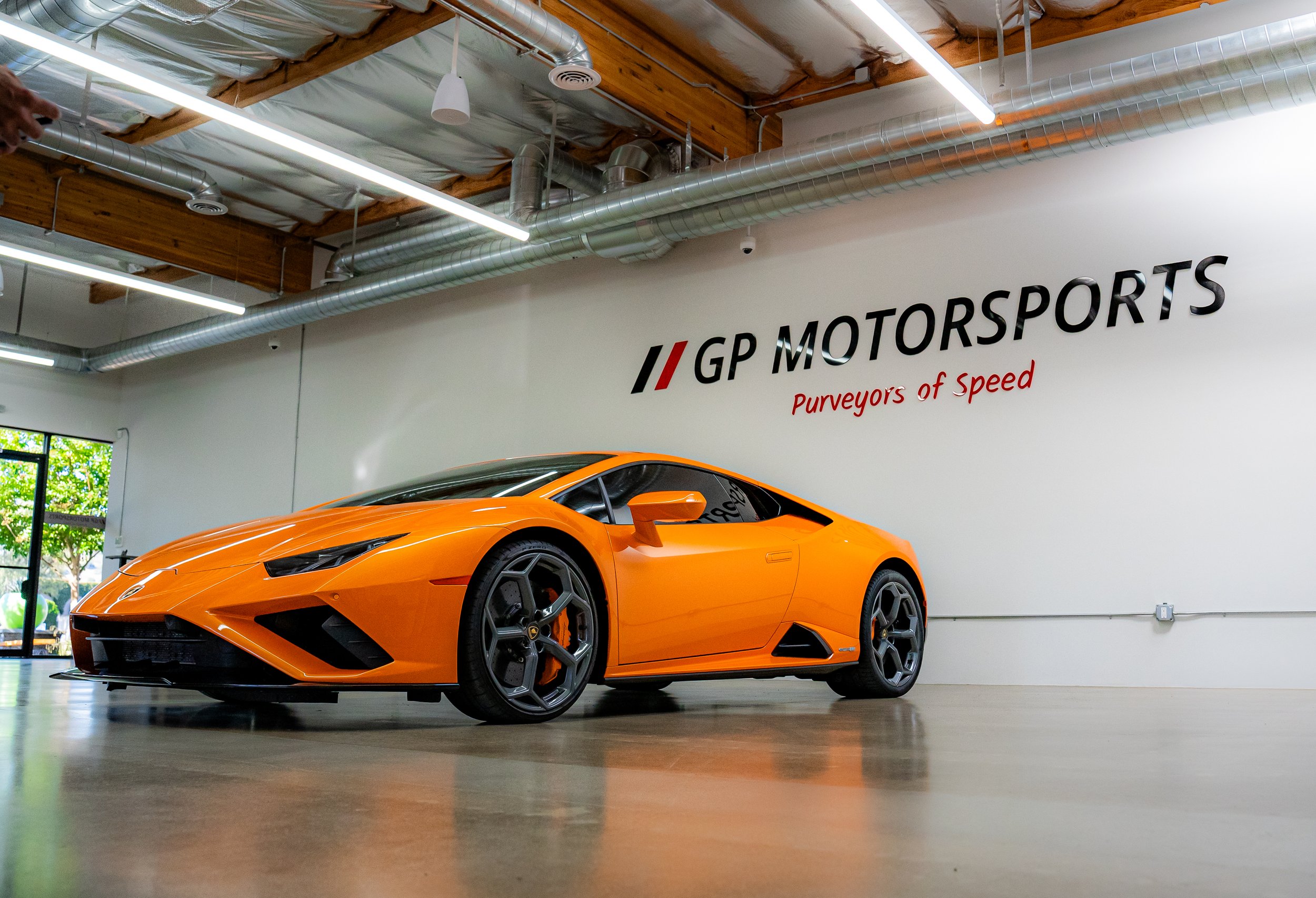 Orange Lamborghini sports car inside a garage with white walls and wooden ceiling beams. The wall features a logo for GP Motorsports and the tagline 'Purveyors of Speed'.
