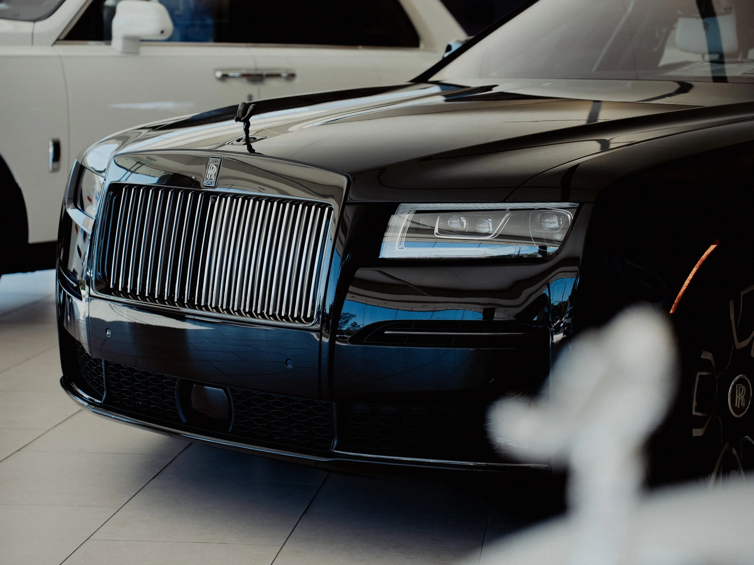Close-up of a black luxury Rolls Royce car's front grille and headlights, with a white vintage car in the background, inside a showroom.