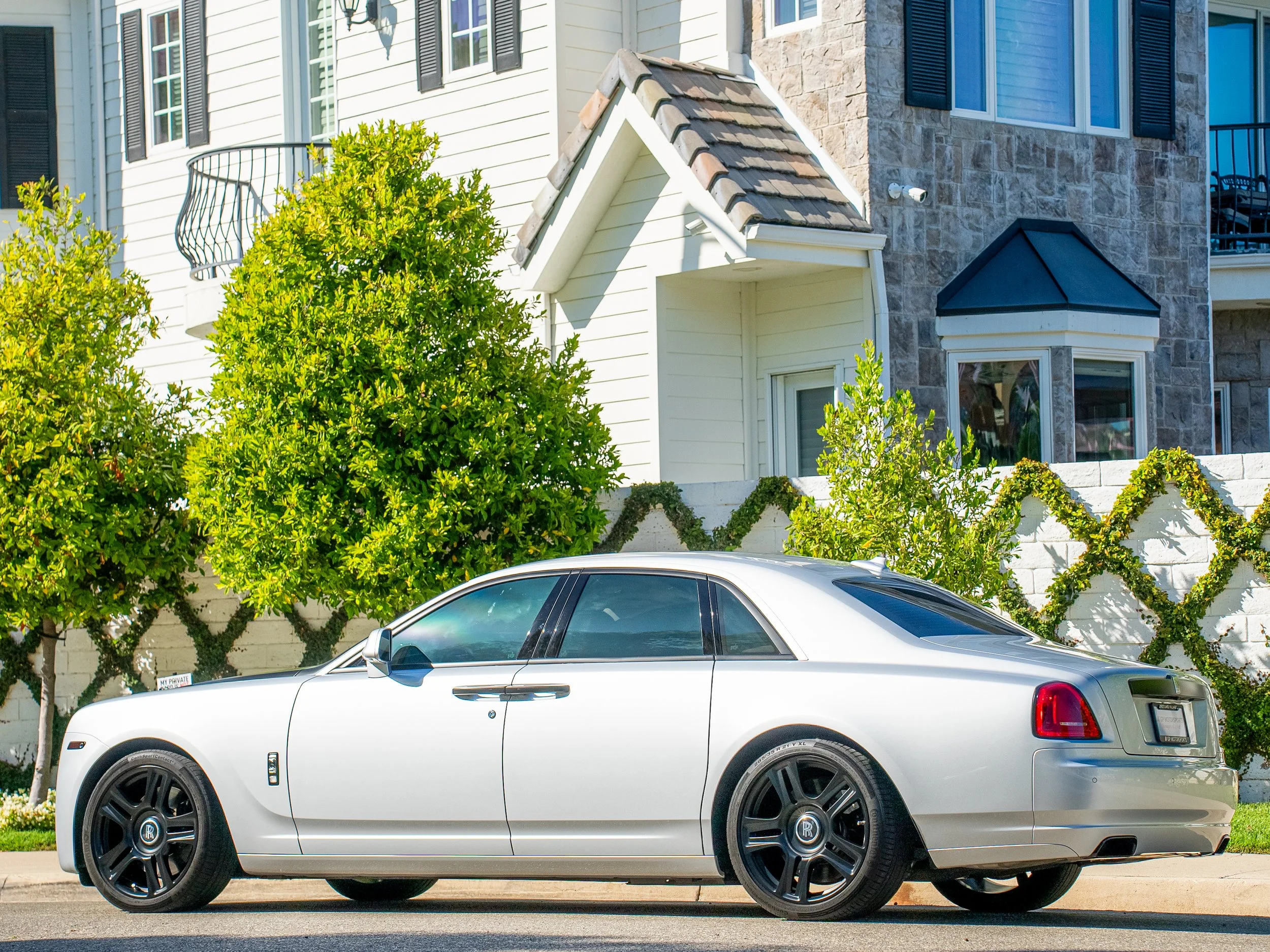 A silver luxury car parked on the street in front of a house with white siding, greenery, and stone accents.