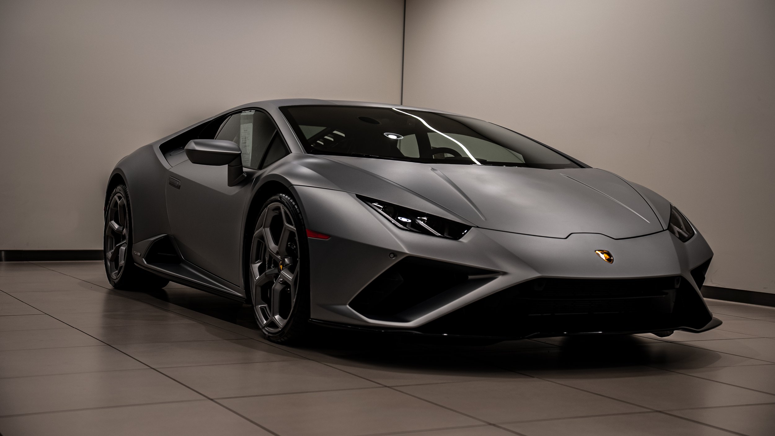 A gray Lamborghini sports car is displayed indoors on a tiled floor, with a plain light-colored wall behind it.