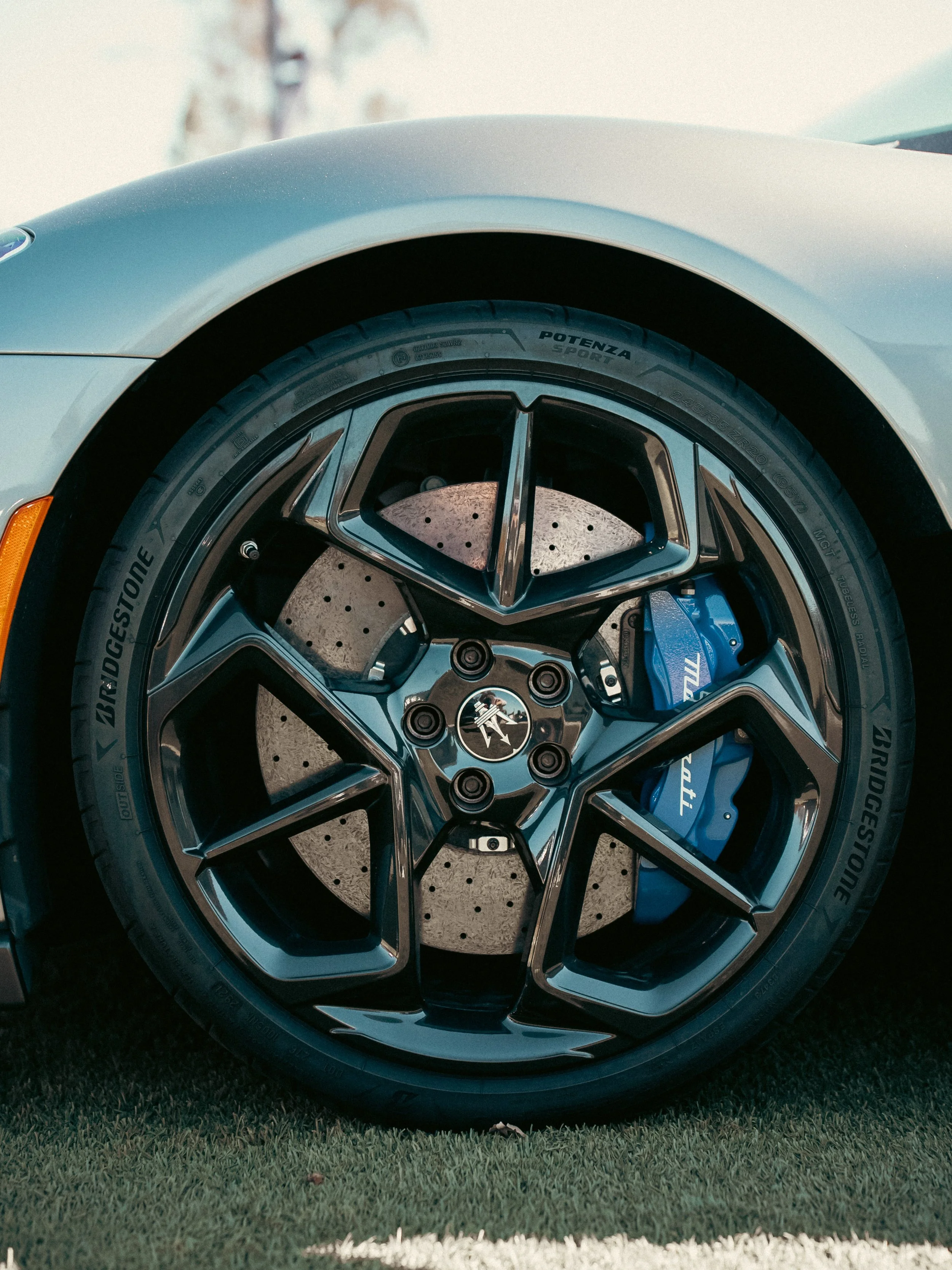 Close-up of a sports car's front wheel showing black rims, a Bridgestone Potenza tire, a large drilled brake disc, and a blue brake caliper.