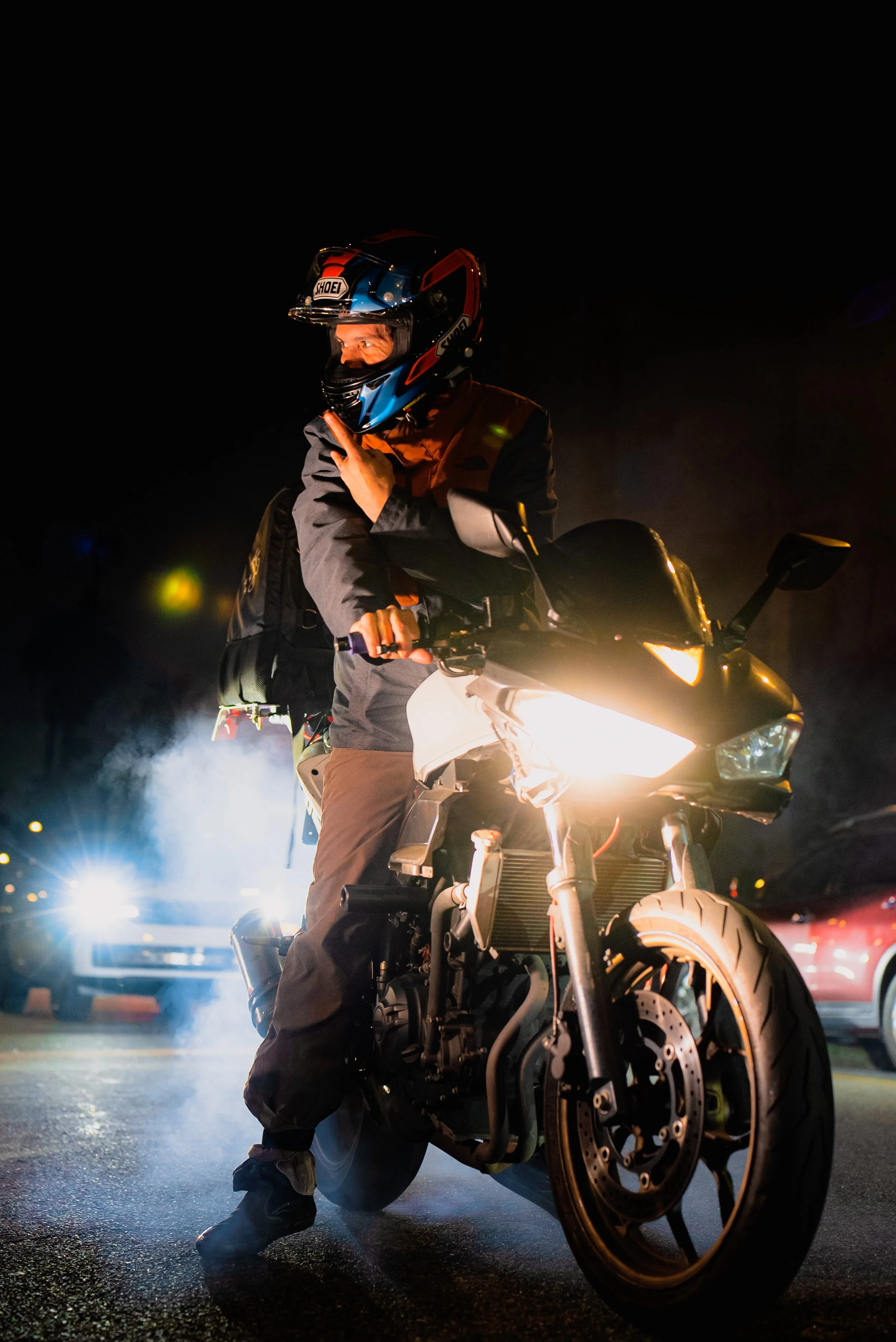 Motorcycle rider at night wearing a helmet, making a peace sign, with headlights on and cars in background