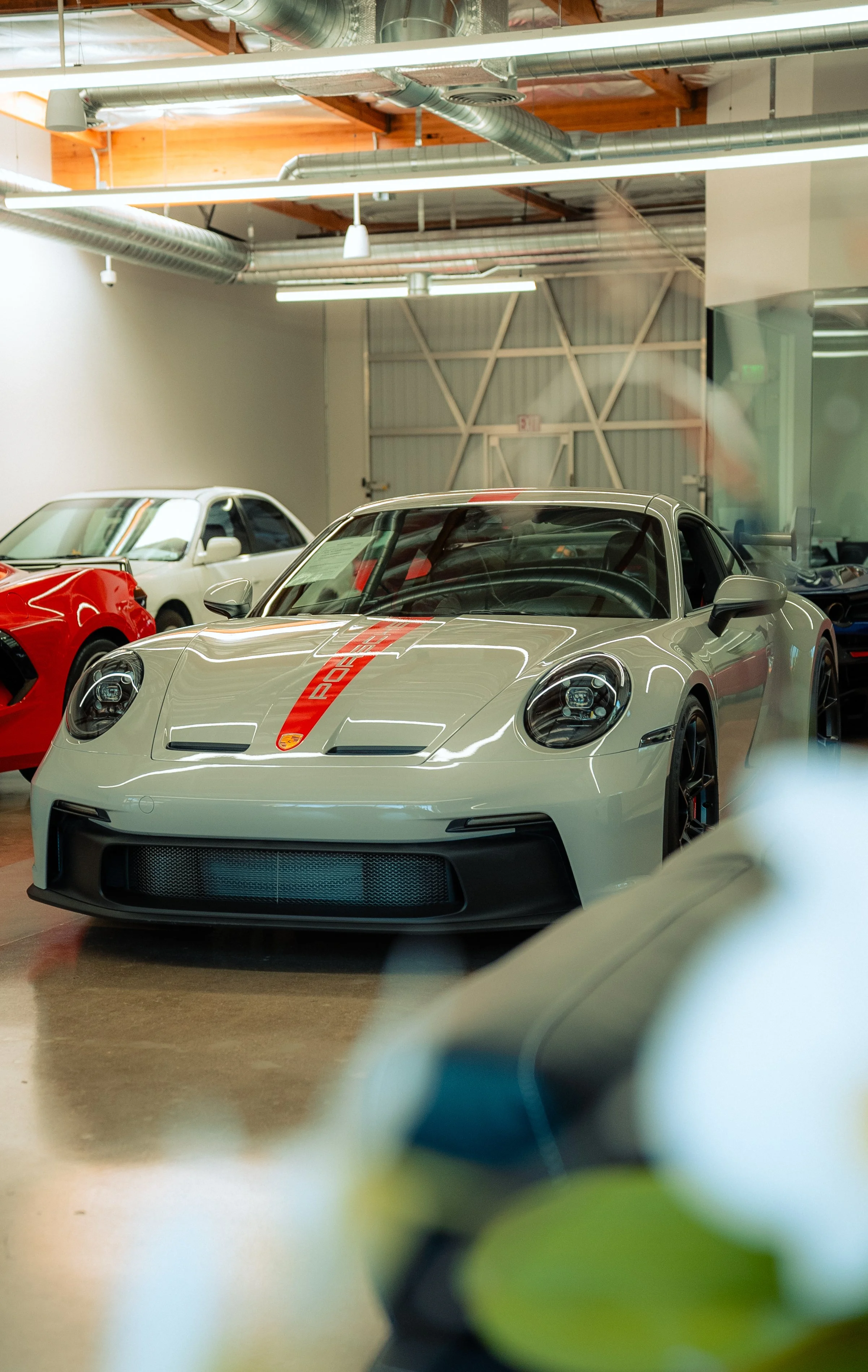 A white Porsche sports car with a red racing stripe down the middle is parked inside a showroom, surrounded by other cars, including a red vehicle and a white car in the background. The showroom has industrial-style exposed pipes and a metal wall.