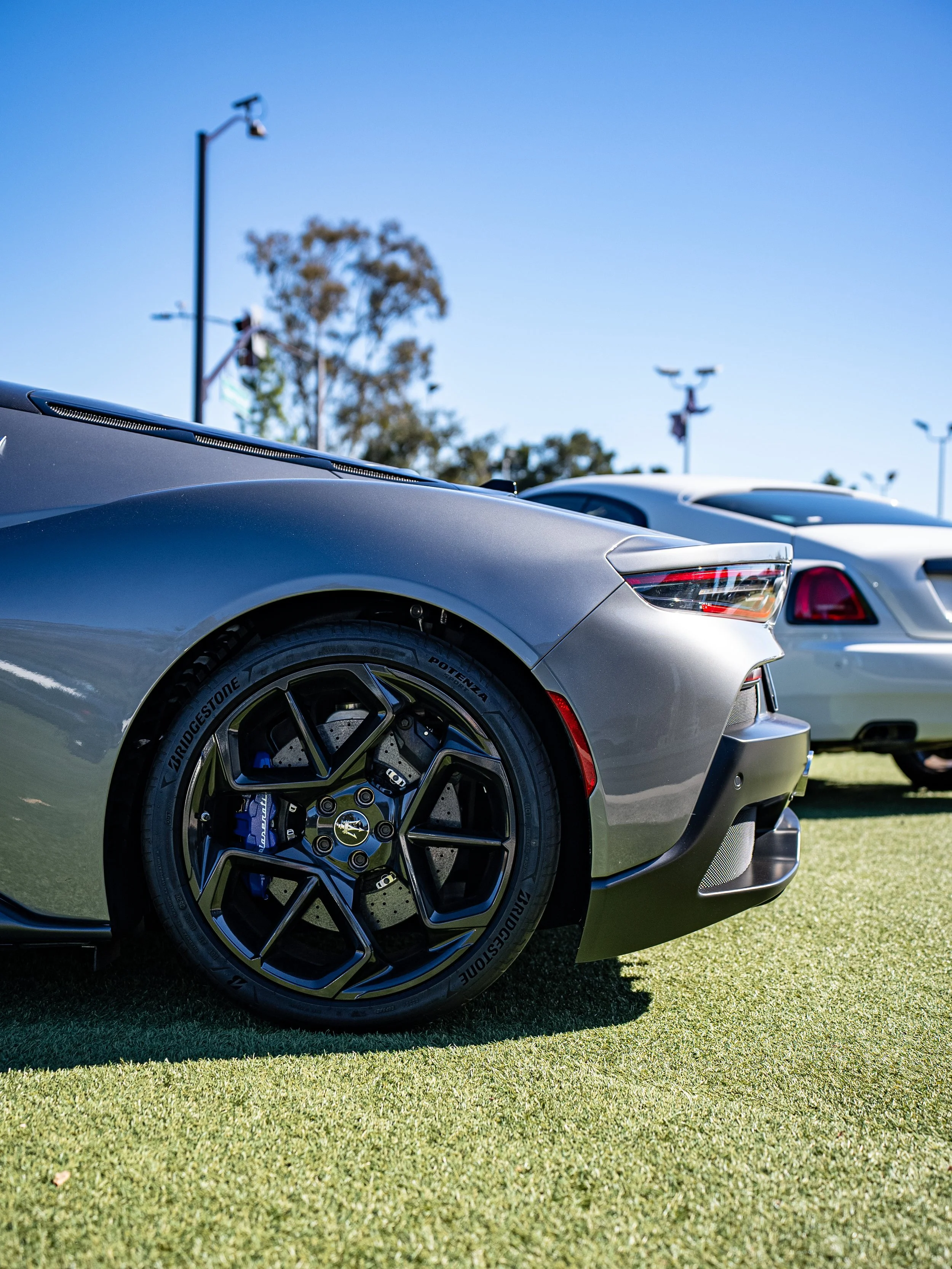 Close-up of a gray sports car with black wheels parked on grass, with other cars and trees in background under a clear blue sky.