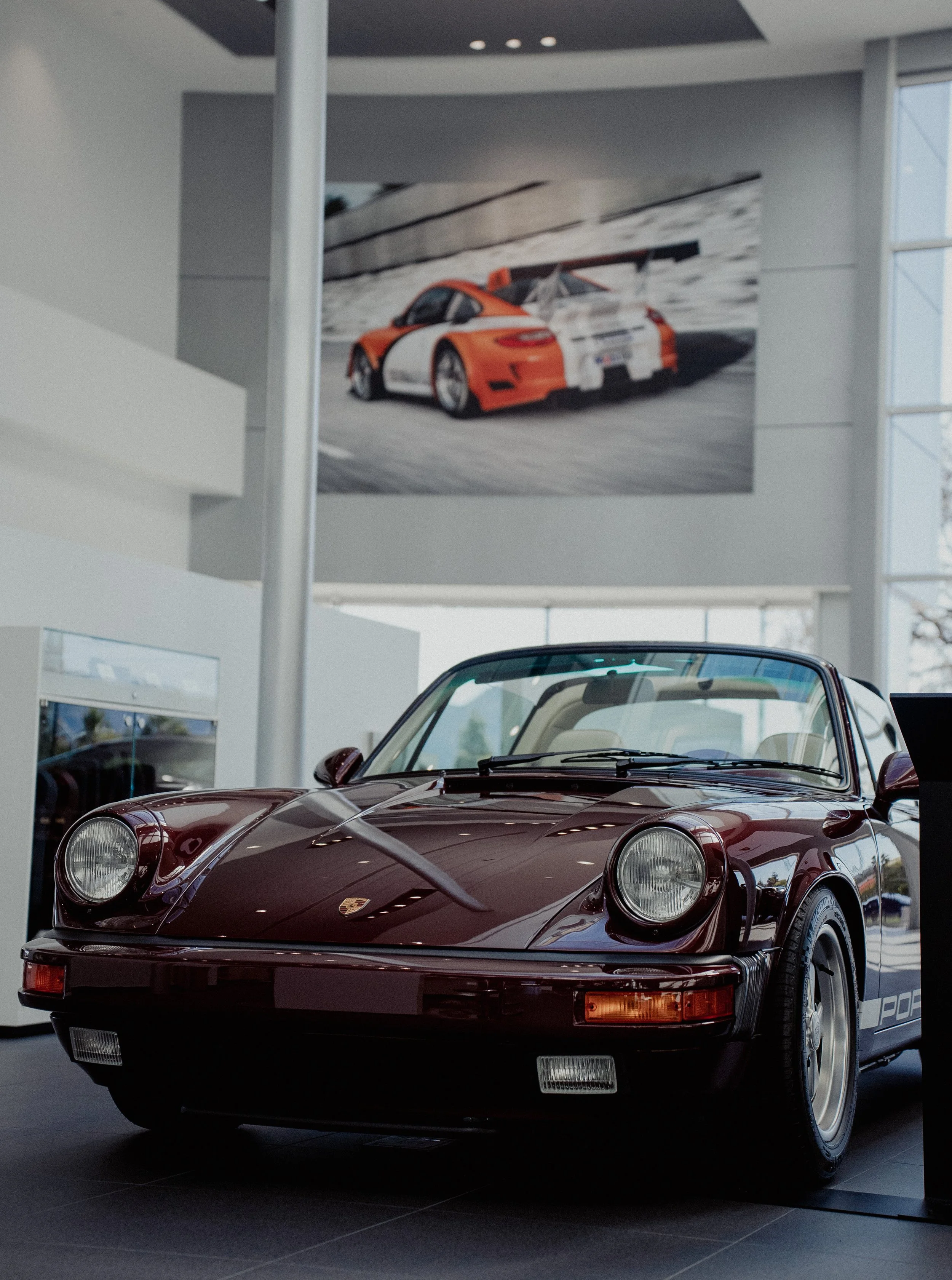 A classic Porsche 911 car on display in a showroom, with a poster of an orange racing version of the same car in the background.