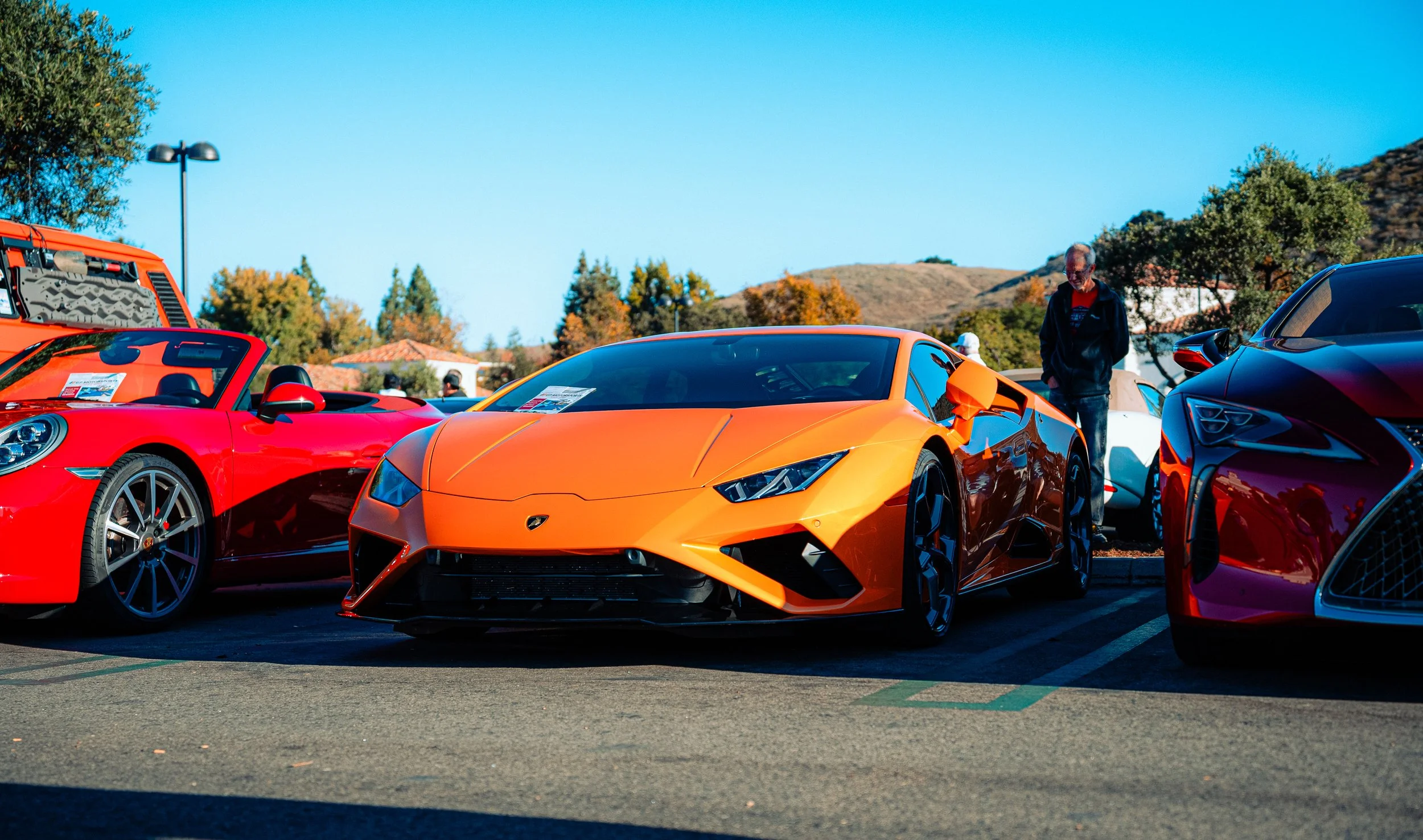 Orange Lamborghini sports car parked between a red Porsche convertible and a red Lexus, with a man looking at the Lamborghini at a car show on a bright day with trees and hills in the background.