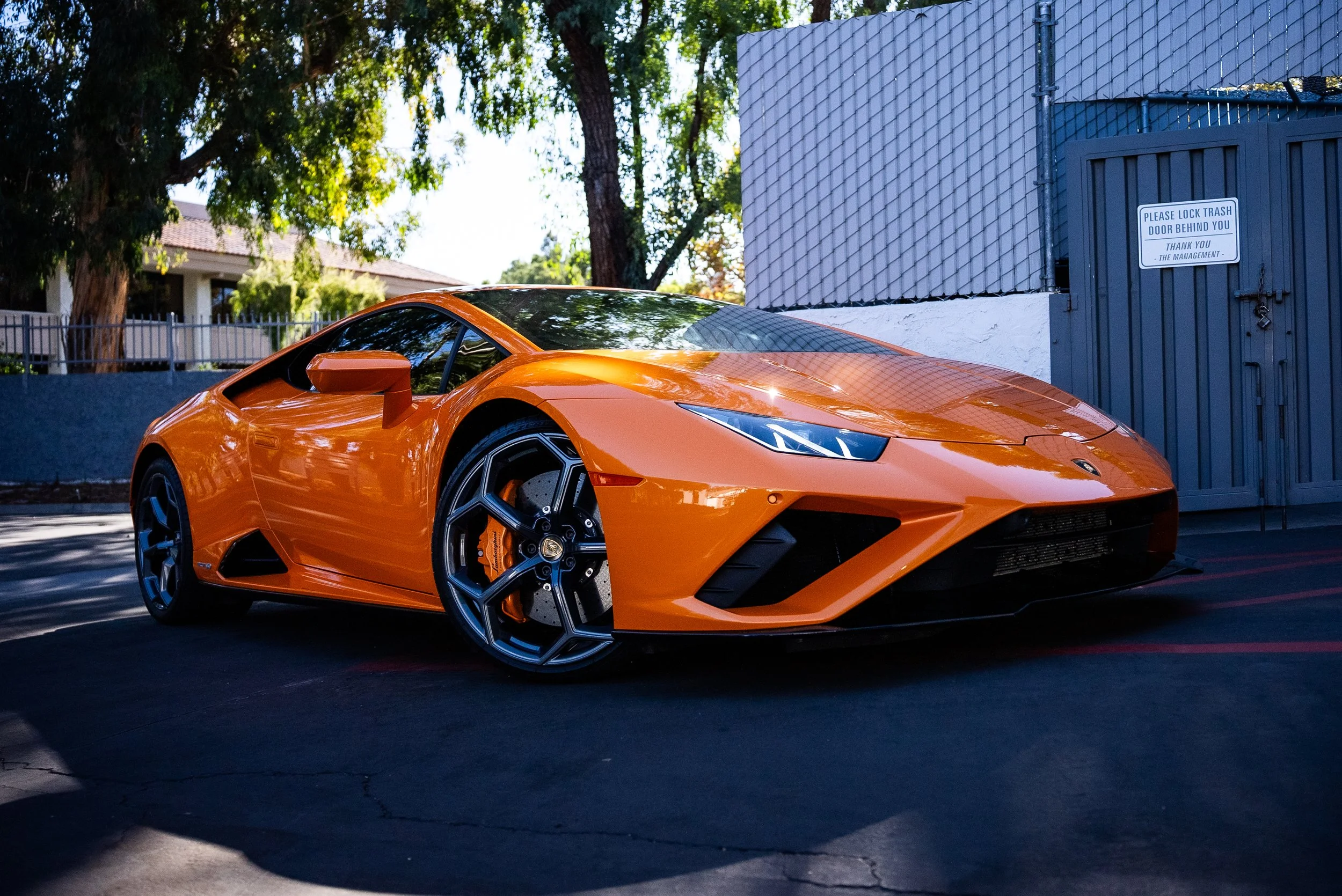 Orange Lamborghini sports car parked in a lot with trees and a blue fence in the background.