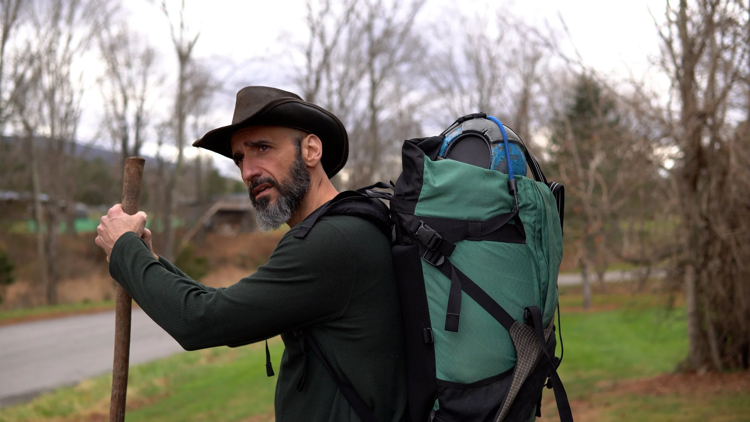 A man standing outside of an old farmhouse, wearing a large pack and carrying a stick